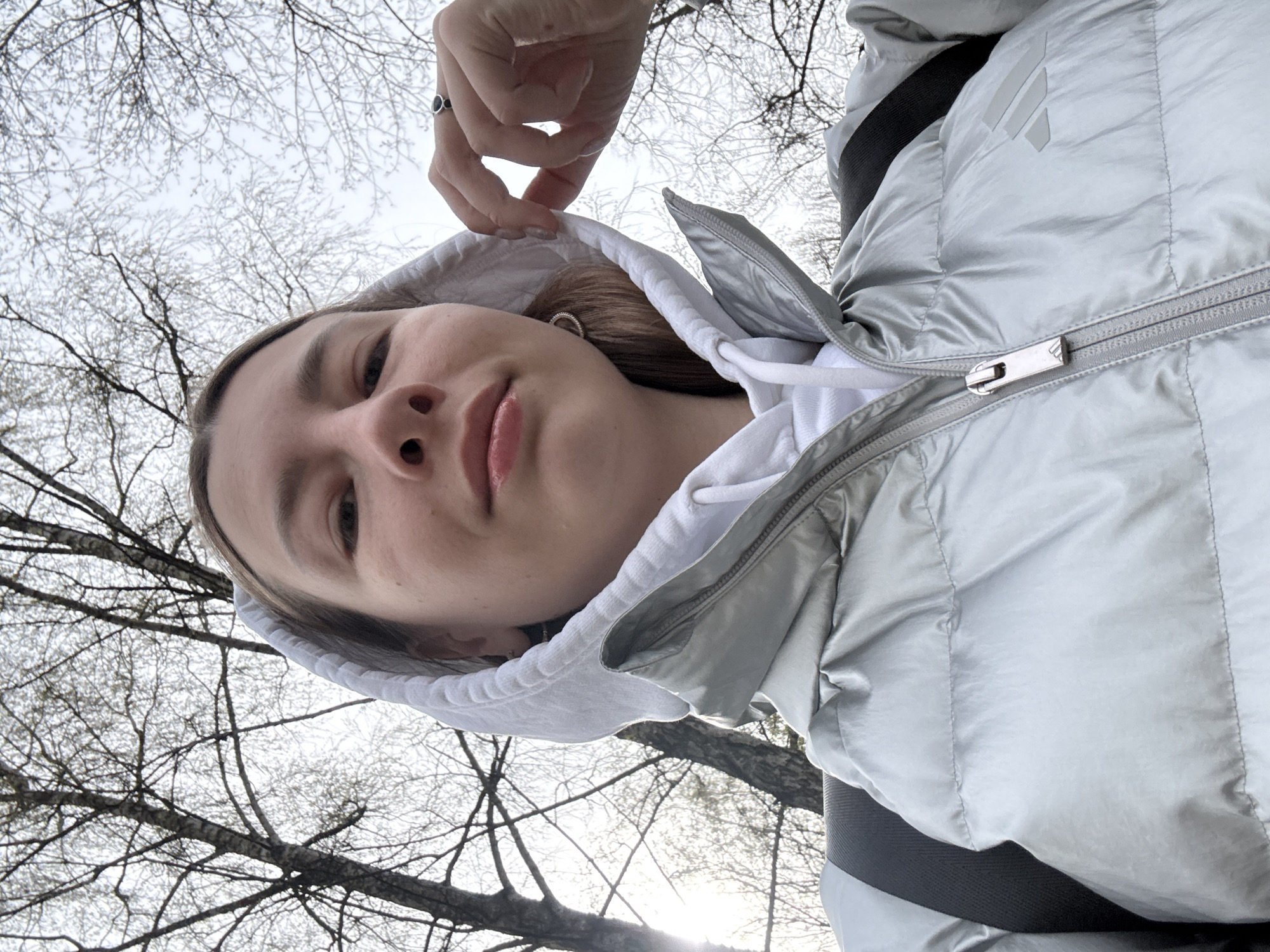 A young woman is captured in a low-angle outdoor shot, looking up towards the camera. Her head and upper torso dominate the foreground, set against a backdrop of bare tree branches reaching towards a bright, overcast sky. She wears a silver or light grey puffer jacket over a white hoodie, with her right hand gently holding the edge of the hoodie around her face. A ring with a dark stone is visible on her right index finger.
Her expression is neutral and relaxed, with natural-looking lips. The visible parts of her hair are light brown. The trees, devoid of leaves, suggest the season is either late winter or early spring in a location like Moscow, Russia. A subtle lens flare or bright spot of light can be seen through the branches, indicating the presence of the sun, possibly diffused by clouds or positioned behind the tree line.
A notable detail on her puffer jacket is the Adidas logo, composed of three diagonal bars, located on the upper left chest area. The zipper pull also features a small, subtle logo. The image conveys a casual, unposed moment, possibly a selfie, taken during daytime outdoors.