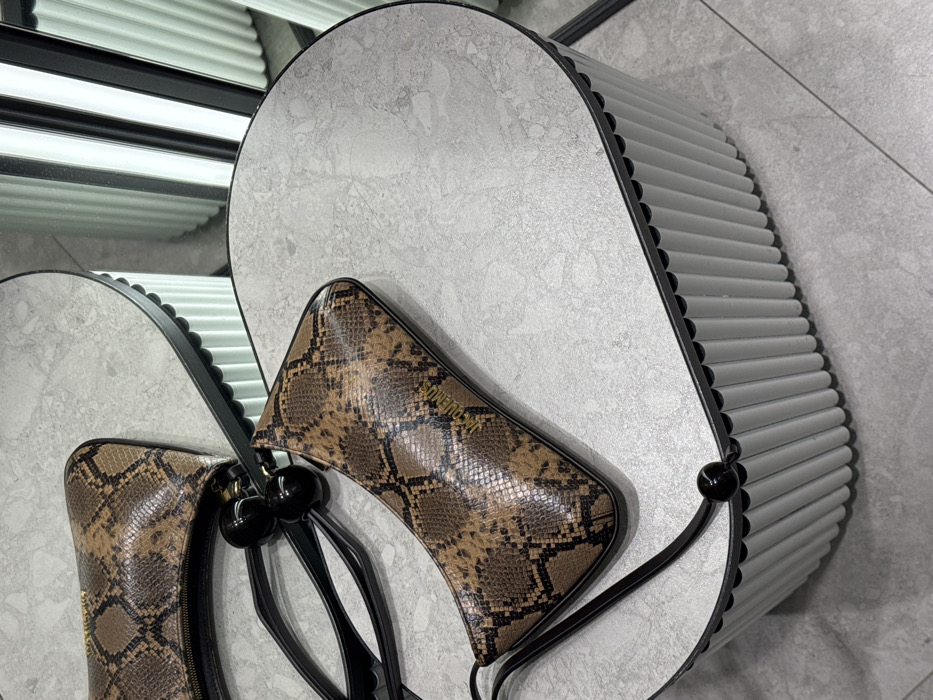 The image displays two Jacquemus handbags, made from brown and black snakeskin-patterned material, positioned on a light grey, mottled, kidney-shaped display table. The table features vertically fluted light grey sides and a black top edge. One bag lies horizontally, partially visible and reflected, while the other is angled diagonally in the foreground, featuring a thin black strap adorned with two prominent black spherical beads. Both bags bear the "JACQUEMUS" logo in gold lettering. The floor consists of large light grey tiles. In the background, a reflection in a mirror reveals vertical, illuminated white panels, suggesting a retail or showroom environment. This scene is located within a luxury department store or high-end boutique, such as TSUM (Central Universal Department Store) in Moscow, Russia.