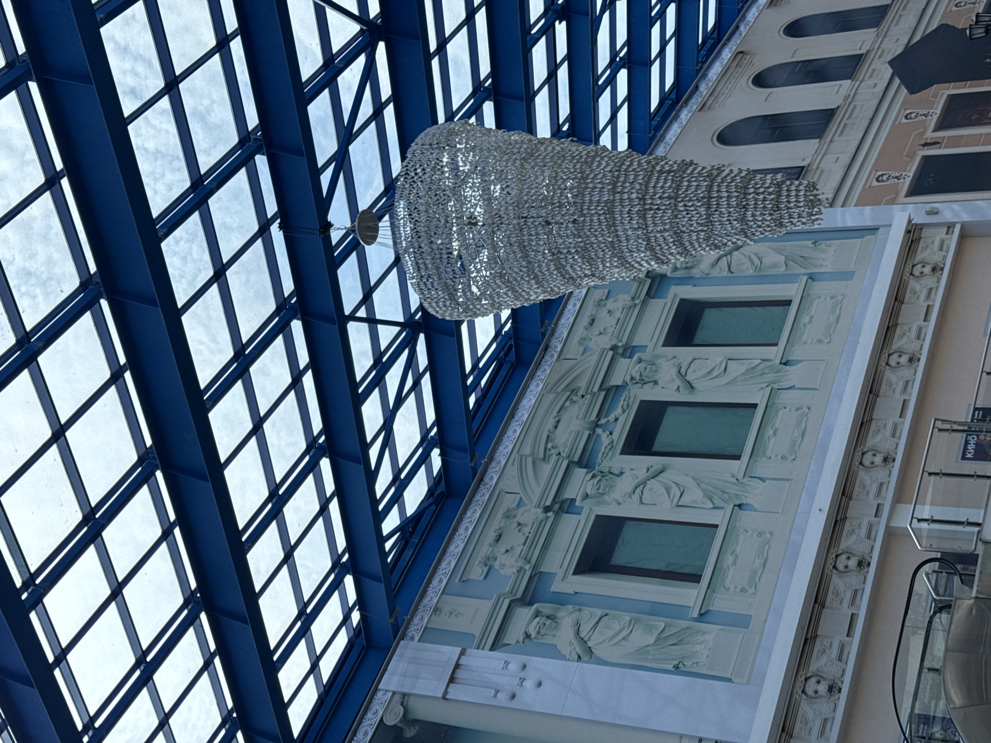 A low-angle shot depicts an interior space, likely a building in Nizhny Novgorod, Russia. A blue grid-like structure of a glass ceiling dominates the left side of the image. Underneath it hangs a large, rounded chandelier of white mesh-like material. Adjacent to the ceiling and chandelier is a cream-colored wall with neoclassical-style decorations, including relief sculptures of female figures under rectangular windows. The wall is decorated with sculptures of heads under the windows. Parts of an escalator are visible on the right, with what appears to be a poster or information board. The orientation of the shot suggests it was taken while standing and looking upward and somewhat toward the right.