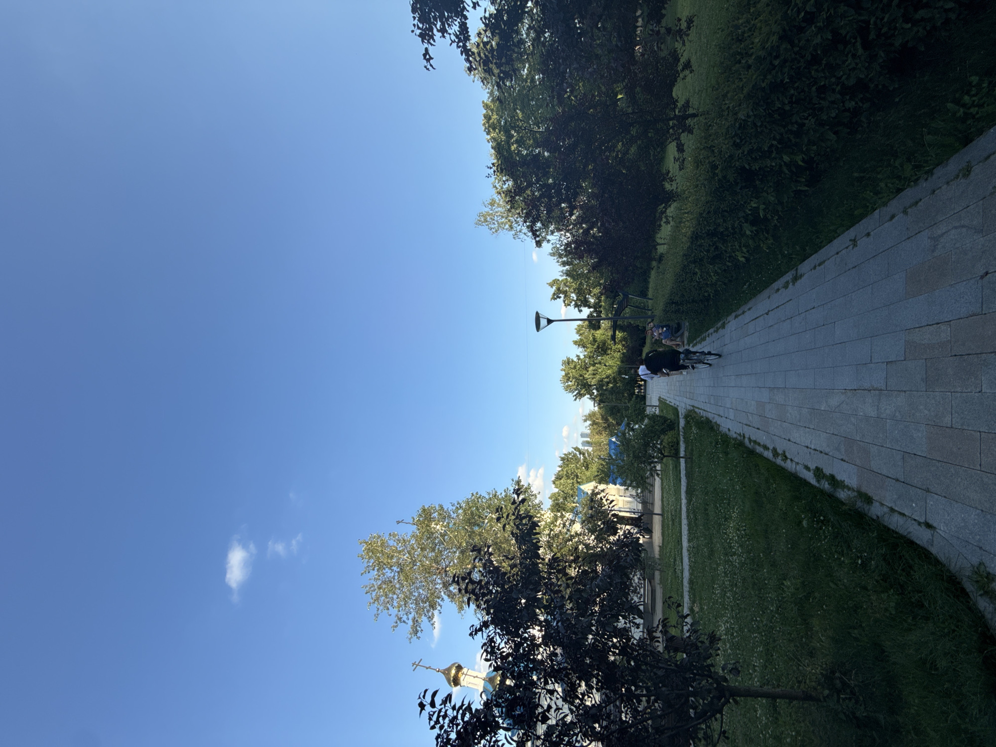 The image depicts a sunlit park path in Moscow, Russia, under a clear blue sky with a few scattered clouds. The path, paved with grey stone tiles, curves to the right, bordered by a lush green lawn on one side and a dense line of trees and bushes on the other.
Several people are visible in the scene. Two individuals are seated on a bench near a lamppost, while another person is walking a bicycle. Further down the path, a group of people are seen in the distance, also near a lamppost.
In the background, through the trees, the golden dome and cross of a Russian Orthodox church are visible, along with some buildings. The presence of the church suggests a location with historical or religious significance.
The overall mood is peaceful and serene, indicative of a pleasant day in the park. The bright sunlight suggests it is daytime, likely mid-morning or afternoon. No visible text is present in the image. The scene captures a typical urban park environment in Moscow during fair weather.
