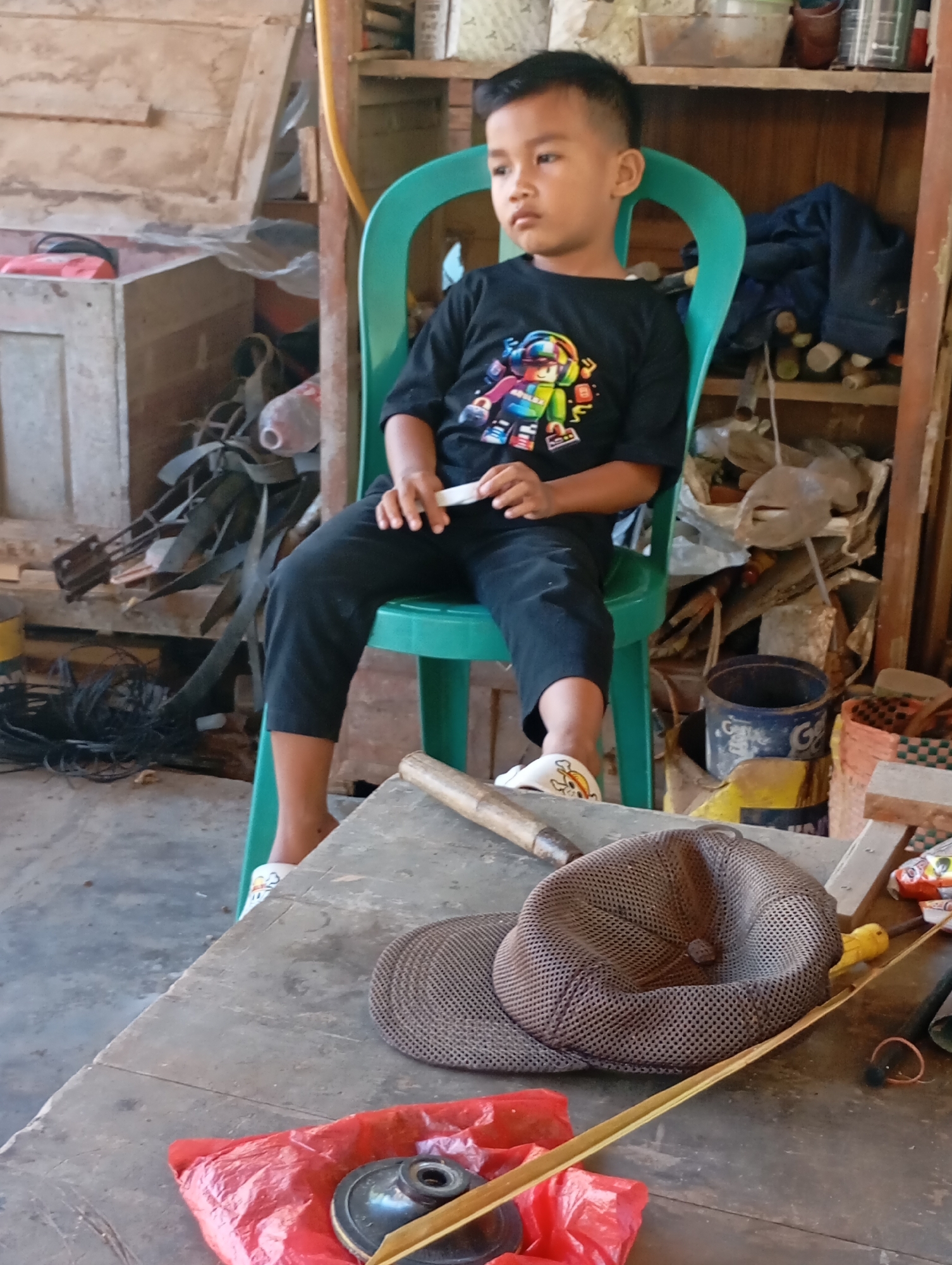 A young boy sits on a bright green plastic chair, his legs crossed at the ankles. He is wearing a black t-shirt with a colorful graphic design and black pants. His hair is neatly cut. The boy looks off to the right, with a neutral expression. In front of him, a wooden table is covered with various objects. A brown mesh cap is placed on the table, next to a piece of wood that appears to be a small bamboo stick. To the left of these items, a red plastic bag contains a dark object, possibly a piece of hardware. The background of the image shows a rustic interior with wooden walls and shelves laden with various items, including bottles, containers, and tools. The scene appears to be in an outdoor workshop or storage area. The location is identified as Ketepung, Indonesia.