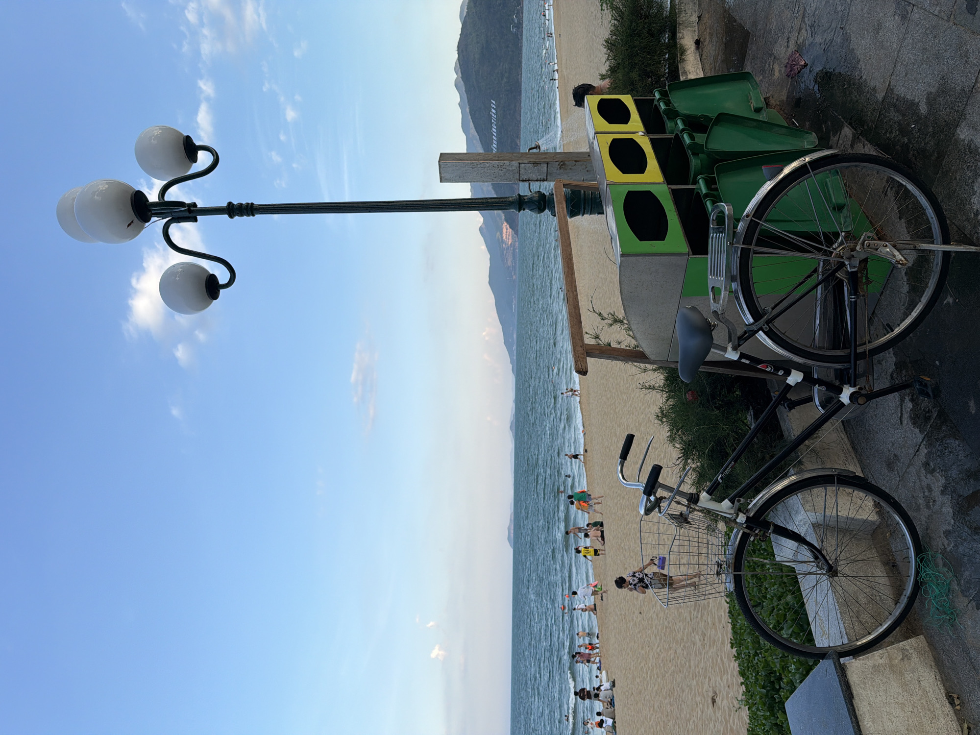 This image depicts a beach scene in Quy Nhon Ward, Vietnam. In the foreground, a black bicycle with a wire basket is parked on a paved walkway. Beside the bicycle are multi-colored recycling bins, one green and yellow, and green bins underneath. A lamppost with three globes stands behind the bicycle and bins.
The sandy beach stretches out to the ocean, where waves are visible. Numerous people are scattered across the beach, some walking, some sitting, and others in the water. The ocean extends to the horizon, with mountains visible in the distance. The sky is blue with a few scattered clouds. The overall impression is a public beach area with recreational activities.