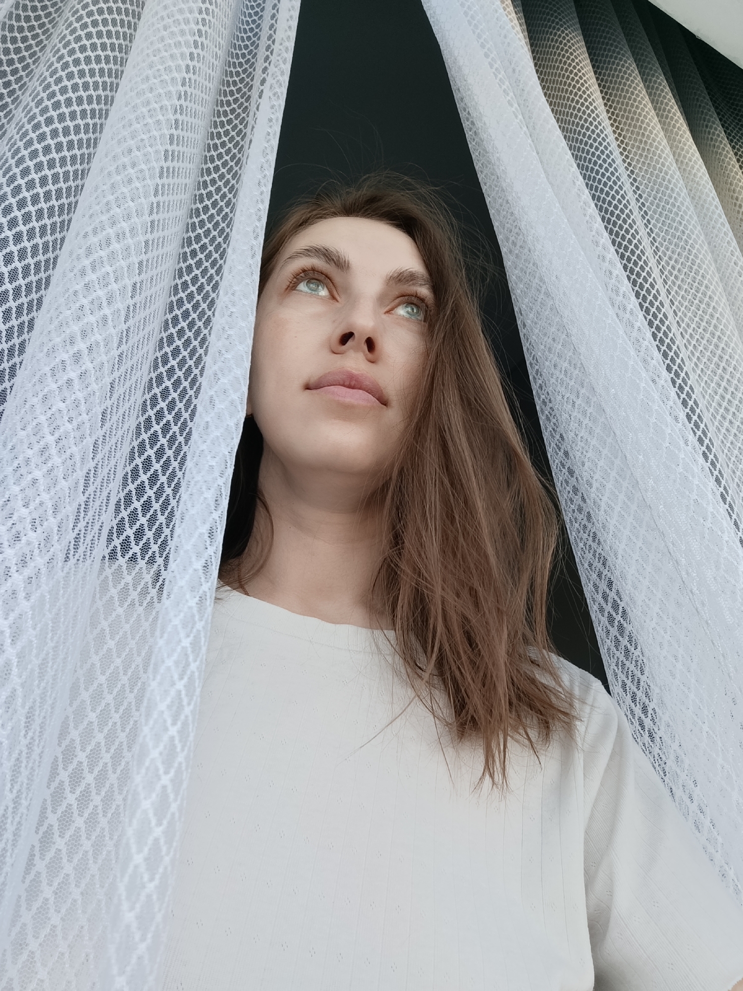 A young woman with long, light brown hair and light green eyes stands indoors, partially obscured by sheer white curtains with a honeycomb pattern. She is wearing a white, long-sleeved shirt. Her gaze is directed upwards and to the left. The location is unspecified, but the request notes the city of Chaikovsky, Russia. The background is dark, possibly a wall or window. The lighting is bright, giving a slightly washed-out appearance to the woman's skin. The setting seems to be a domestic environment near a window.
