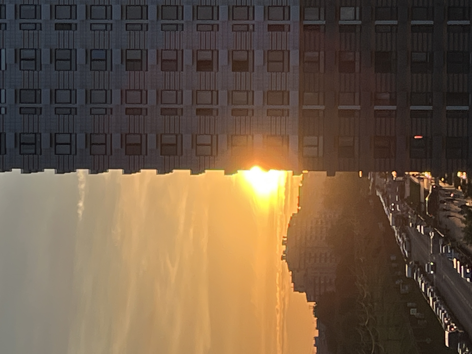 The image depicts a wide-angle view of a city street at sunset, likely in Moscow, Russia. A tall, modern apartment building dominates the right side of the frame, its facade featuring rows of rectangular windows. The setting sun, a bright orange orb, is partially obscured by the building on the left, casting a warm, golden glow across the sky and illuminating a line of clouds.
In the background, other buildings are visible, including what appears to be a large, multi-story residential complex with a distinctive roofline, also bathed in the sunset's light. A tree-lined street or park area separates the apartment building from the background structures, with power lines visible in the mid-ground.
The street in the foreground is densely packed with cars, indicating a traffic jam. The vehicles are mostly sedans and SUVs, with a few vans or trucks interspersed. Their headlights are not yet on, suggesting it is still daylight. The road surface is dark asphalt, and the overall impression is one of urban density and a busy evening commute. No people are clearly visible in the image. The scene is characterized by its warm, atmospheric lighting due to the sunset.