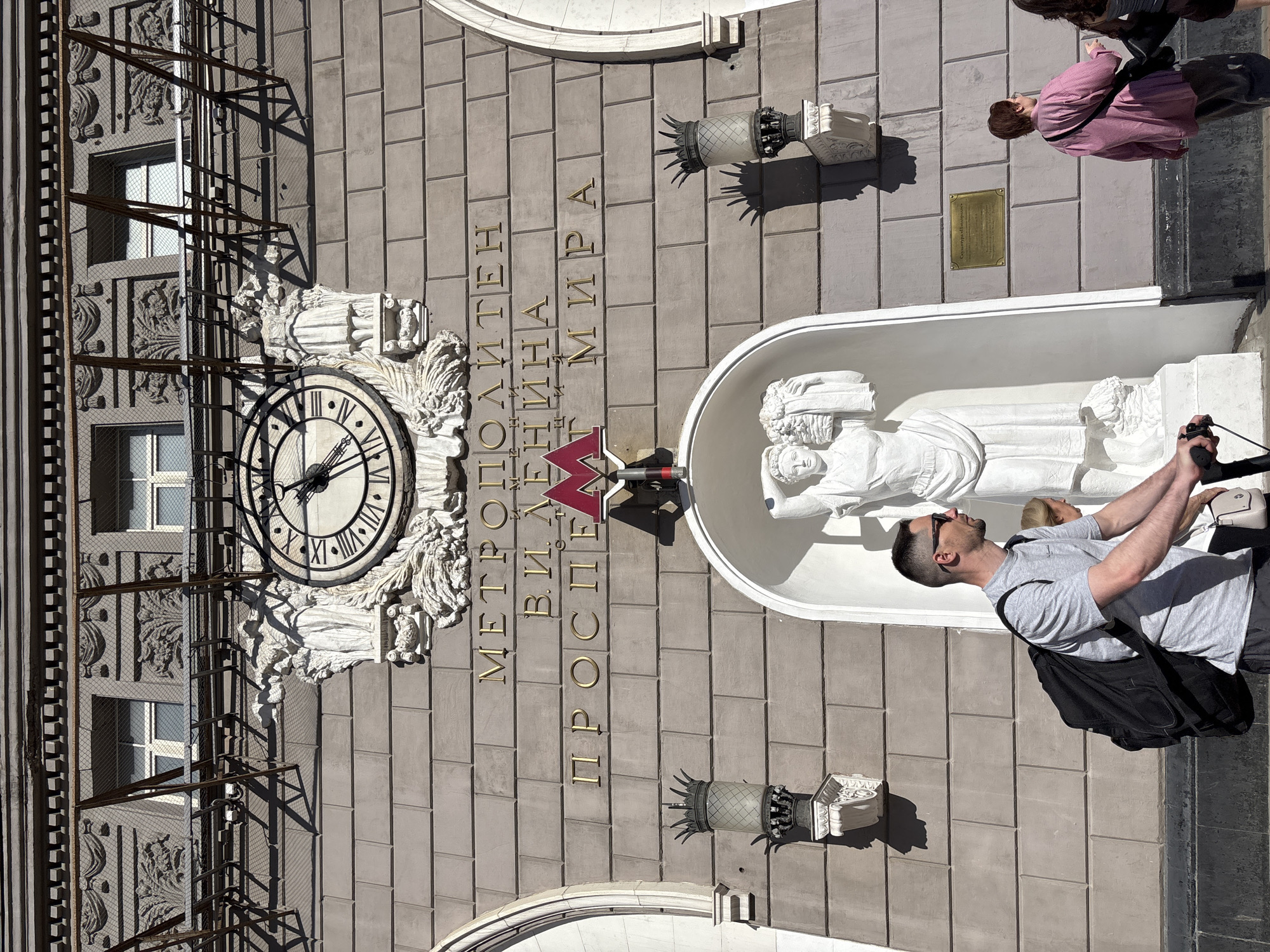 The image shows a frontal view of the Prospekt Mira metro station building in Moscow, Russia. The building's facade is constructed of large, grey stone blocks. A prominent clock face with Roman numerals is mounted high on the facade, above ornate sculpted figures. Below the clock, golden Cyrillic lettering spells out "МЕТРОПОЛИТЕН В. И. ЛЕНИНА ПРОСПЕКТ МИРА" which translates to "LENIN METROPOLITAN PROSPEKT MIRA". A red 'M' logo, the symbol for the Moscow Metro, is positioned centrally below the lettering. Two decorative lampposts with intricate metalwork are attached to the wall on either side of the entrance.
In the foreground, a man wearing sunglasses and a grey t-shirt with a black backpack is riding a scooter. He is moving from left to right across the frame. Partially visible behind him, a woman with blonde hair is also walking. To the right, another woman, wearing a pink and white striped shirt and dark trousers, walks away from the camera. In the lower right corner, another person's leg and hand holding the scooter handle are visible.
A large white sculpture of a woman holding a bouquet of flowers is housed in an alcove beneath the station's lettering. The sunlight casts strong shadows across the building and the figures.