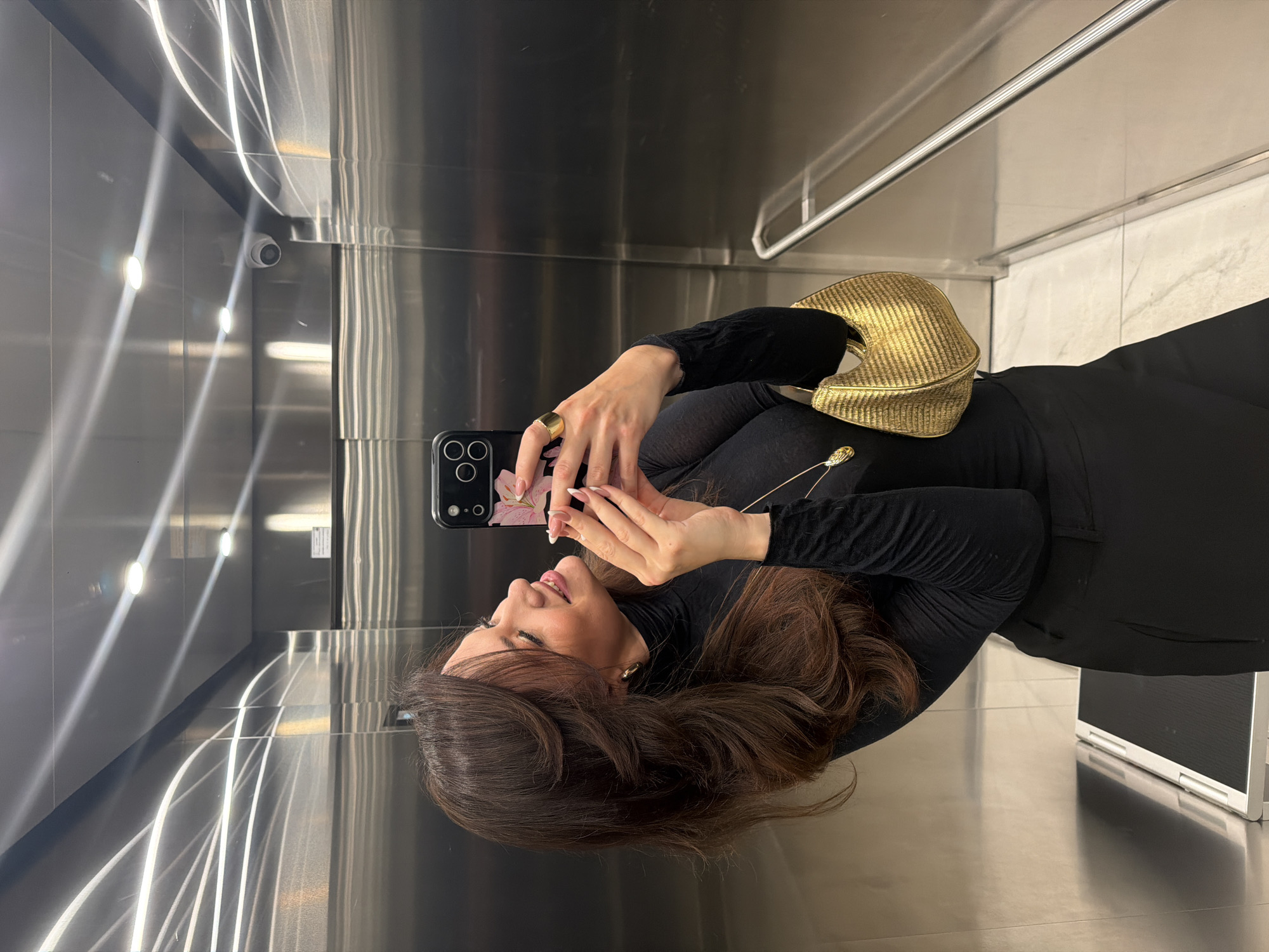 A woman is taking a selfie with a smartphone inside a modern elevator. The elevator has stainless steel walls and ceiling, reflecting the light from ceiling fixtures. The woman has long brown hair and is wearing a black turtleneck and pants. She has a gold ring on her finger and a gold purse over her shoulder. A security camera is mounted on the elevator wall. Based on the architectural style of the elevator interior, it is likely located in a modern building in Belgrade, Serbia.