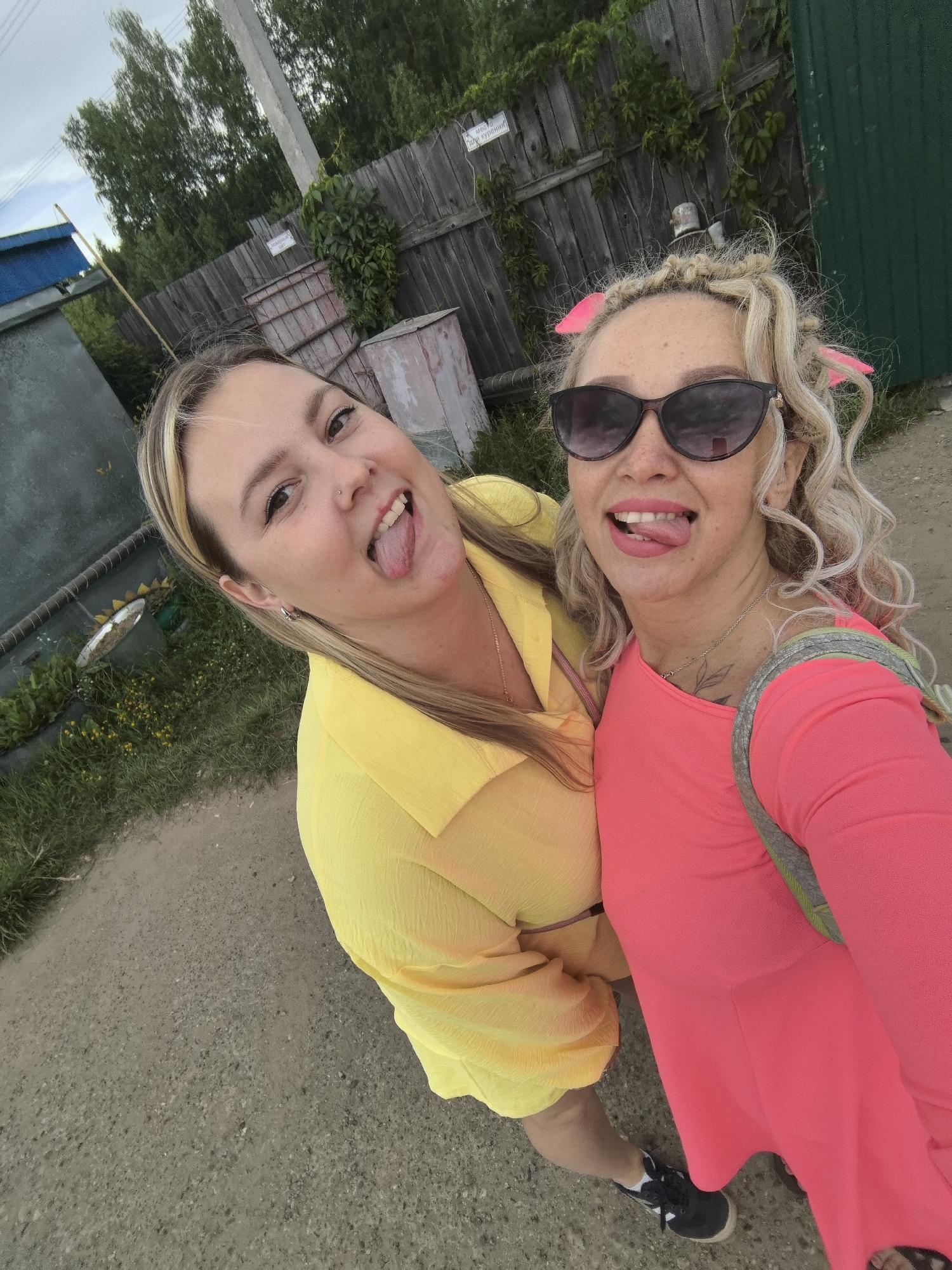 Two women are taking a selfie outdoors, possibly in a rural setting in Noviy, Russia. They are standing on a dirt or gravel path. The woman on the left is wearing a bright yellow shirt and has blonde hair. She is sticking her tongue out and smiling. The woman on the right has curly blonde hair with pink accessories and is wearing a bright pink dress and sunglasses. She is also sticking her tongue out and smiling. Behind them, there is a wooden fence covered in vines, some greenery, and what appears to be industrial or agricultural structures. A tall, slender pole, likely a utility pole, is visible to the left of the fence. The sky is overcast, suggesting it might be a cloudy day. The overall mood of the image is playful and happy, with both women expressing a sense of fun and camaraderie. Visible text on a white sign attached to the fence reads "МОСТО" and another sign below it has some Cyrillic text that is partially obscured but appears to be "КУРЕНИЯ".