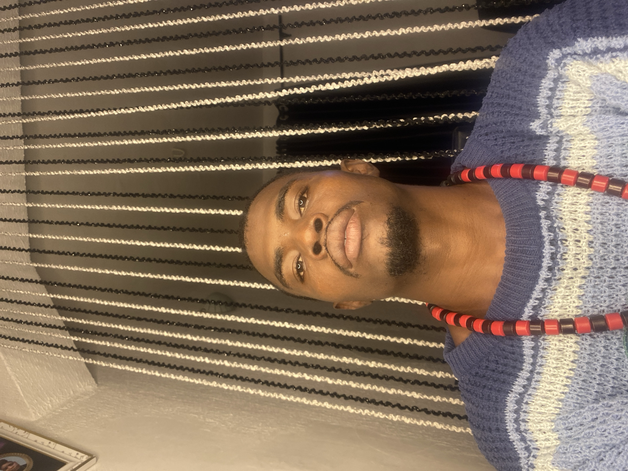 A medium shot captures a Black man from the chest up, looking directly at the camera. He is wearing a blue, white, and light blue striped knitted sweater and a necklace made of red and black beads. His head is slightly tilted back, and he has a goatee. The background consists of vertically hanging strands of black and white beads, creating a textured curtain effect. To the left of the man, a framed picture is partially visible on a textured white wall. The lighting is somewhat dim, casting soft shadows on his face and the background. This scene appears to be indoors, likely in a residential setting within Abakaliki, Nigeria.