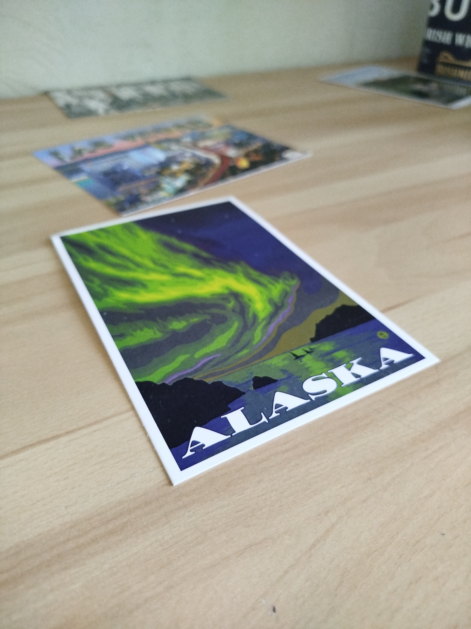 A high-angle, close-up shot shows a postcard of an Alaska landscape resting on a wooden surface. The postcard depicts an aurora borealis in shades of green and yellow against a dark blue sky, with silhouettes of mountains and water below. "ALASKA" is printed in large, white, capitalized letters at the bottom. Other postcards are visible in the blurred background, along with a dark object in the top right corner. The setting does not appear to be in Varna, Bulgaria; it is a tabletop indoors.