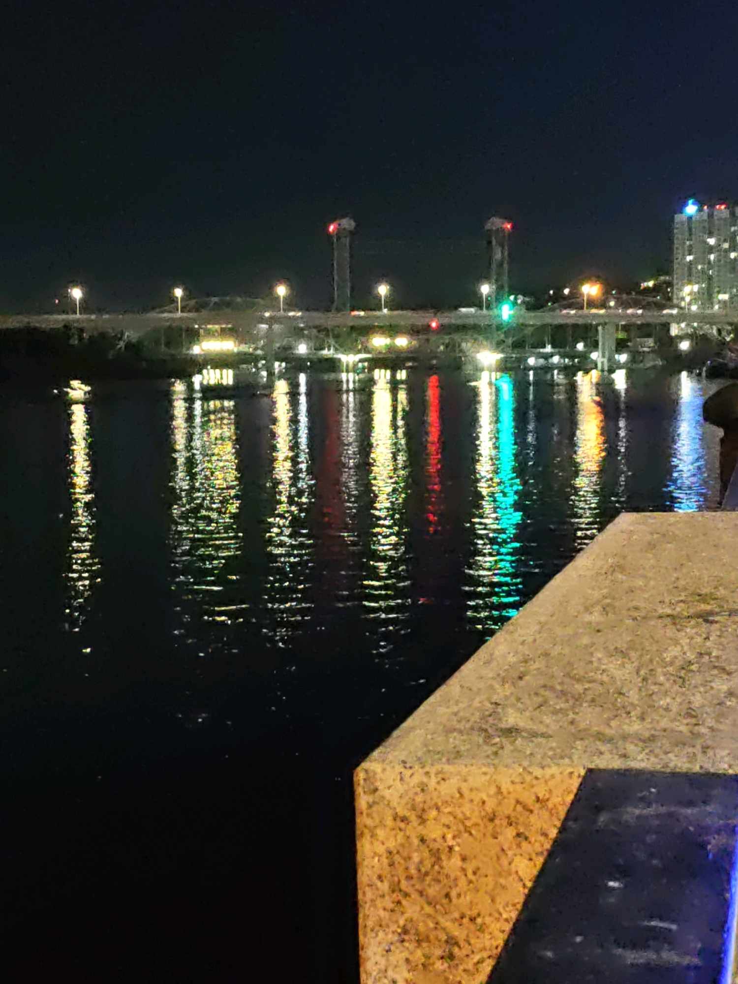 The image captures a night view of the Don River in Rostov-on-Don, Russia, with the Voroshilovgradsky Bridge illuminated in the background. The bridge, a prominent landmark, features several illuminated pylons and traffic lights, casting colorful reflections onto the dark water. The reflections are a mixture of red, green, yellow, and blue lights, creating a vibrant and abstract pattern on the river's surface. On the right side of the frame, a portion of a concrete embankment is visible in the foreground, with a dark blue railing. The sky is dark, suggesting late evening or night, with minimal visible stars. A tall, lit building can be seen in the distance on the right, adding to the urban cityscape. The overall scene depicts a lively urban waterfront at night, characterized by artificial light and its reflection on the water.