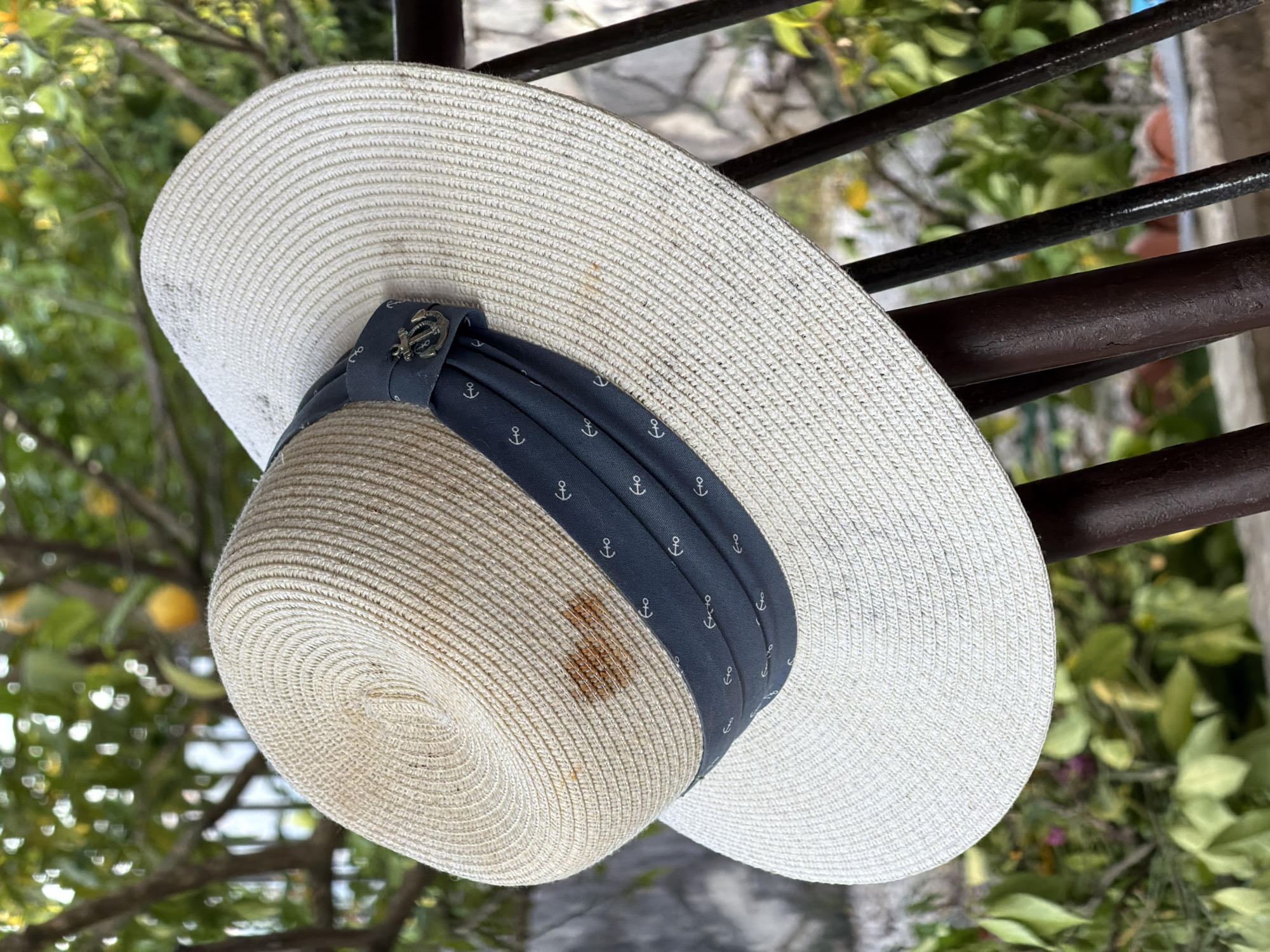 A straw sun hat with a navy-blue ribbon adorned with anchor symbols hangs on a dark brown metal railing. The hat has a wide brim and is off-white with some visible stains. The railing is composed of several horizontal bars. The background reveals a blurry landscape featuring greenery and hints of a building. The scene is located in Gošići, Montenegro.