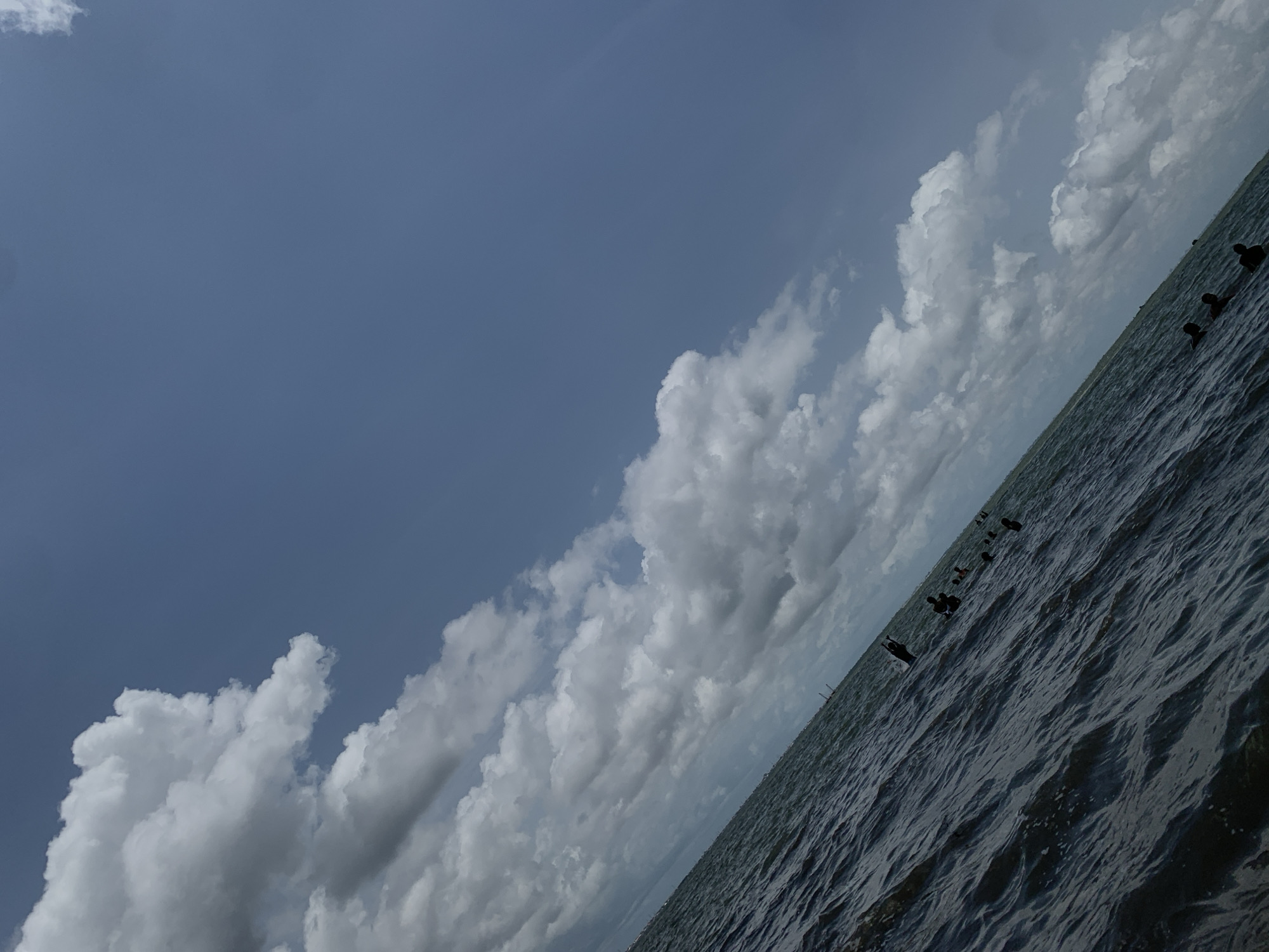 The image depicts a body of water under a partly cloudy sky, likely taken in Ikorodu, Nigeria. The water surface is textured with ripples, suggesting a slight breeze. Dark shapes, possibly birds or other floating objects, are scattered on the water surface. The sky is predominantly blue with patches of white clouds, ranging from fluffy cumulus to layered stratus formations. The horizon line is not distinct, as the water blends into the distant sky.