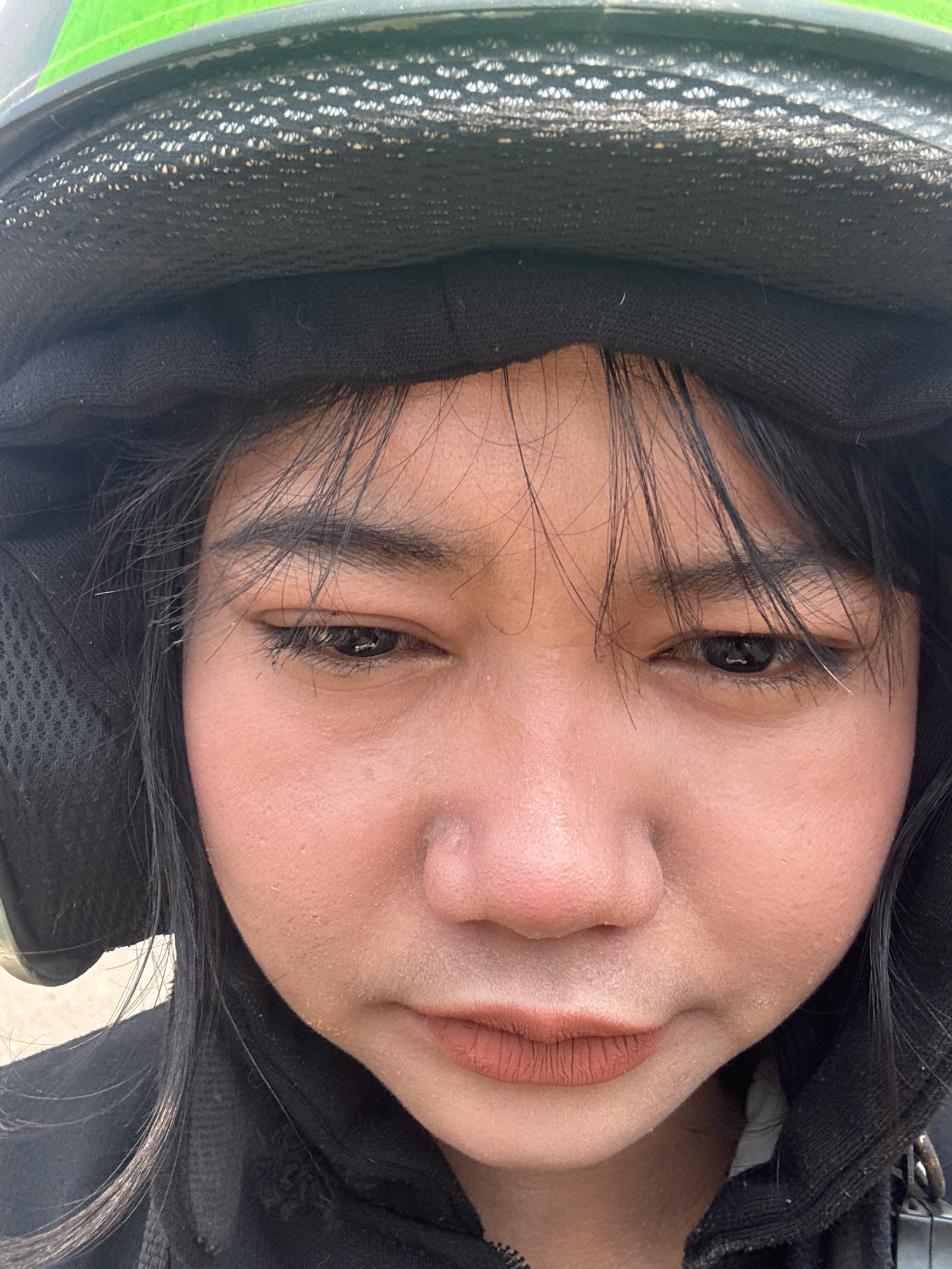 The image is a close-up, front-facing selfie of a young woman wearing a green and black helmet and a black hooded jacket. She has black hair that falls around her face, and her eyes are visible. Her lips are colored with a matte brown lipstick. The background is out of focus, but appears to be outdoors. The lighting suggests it is daytime. There are no other people or significant objects in the frame besides the woman and her attire. No specific location in North Jakarta, Indonesia is discernible from the image itself.