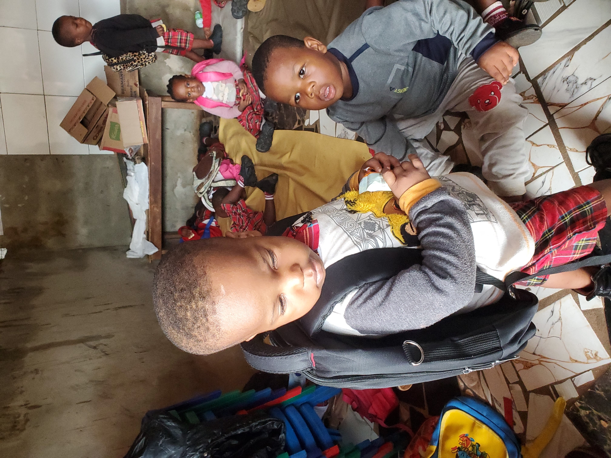 This image captures a group of young children indoors in what appears to be Ukwu-Abwa, Nigeria. The photograph is taken from a slightly elevated perspective, with the children mostly on the floor.
In the foreground, a young boy with closely cropped hair is seen lying on his side, his head resting on what appears to be a black backpack with silver rings. He is wearing a grey long-sleeved shirt and a red and white plaid bottom, and his eyes are partially closed, giving a peaceful or sleepy expression. He has a yellow plush toy, possibly a giraffe, near his hands.
Dominating the mid-ground is another young boy, wearing a grey long-sleeved shirt with a dark blue accent and the partially visible word "ENERGY" on it. He looks directly at the camera with a curious expression and slightly open mouth. He is either sitting or kneeling on a yellow fabric, and his left leg shows a white and red striped sock and a light grey pant with a small red bear emblem. His right hand holds what looks like a small wrapped item, possibly money.
Further back and to the left, several other children are visible. One young girl wears a pink jacket and a plaid skirt, looking towards the camera. Another boy, standing, wears a black top and plaid shorts, with a leopard print backpack on his back. Other children are partially visible in the background, many clad in plaid patterns or bright colors.
The setting appears to be a multi-surfaced indoor space. The foreground and mid-ground show a tiled floor with white and dark, marbled patterns. To the left, there's a darker, rougher concrete floor. Scattered around are various objects: cardboard boxes (one with the word "BENMAN" partially visible), colorful blocks or toys in shades of blue, green, and red, a black plastic bag, and a small blue and yellow backpack featuring a cartoon character, possibly from Teenage Mutant Ninja Turtles. The overall scene suggests a vibrant, active environment, likely a community space or daycare. The time of day is bright, consistent with indoor daylight.