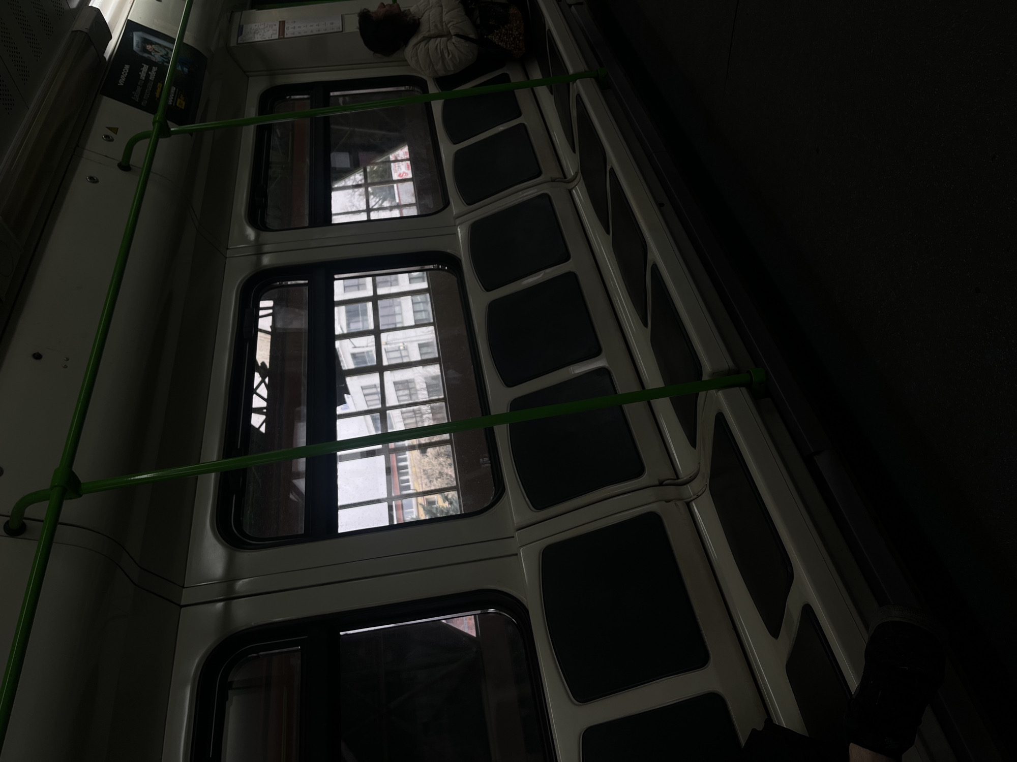 The image depicts the interior of a metro train in Sofia, Bulgaria. The perspective is from a lower angle, looking across a row of empty passenger seats towards the windows. The train car features white wall panels and light-colored seats with dark, rectangular cushions. Green metal grab bars extend horizontally across the upper section of the frame and vertically towards the floor. Through the multiple windows on the right, an urban exterior is visible, consisting of multi-story buildings with numerous windows, bare tree branches, and a distant red sign with white lettering. Above the windows, a "VIVACOM" advertisement with Cyrillic text is mounted on the wall. On the far right, an adult individual, appearing to be female, is seated, wearing a light-colored (possibly white or beige) puffer jacket and holding a patterned bag. Her head is turned towards the right, mostly out of frame. In the lower left corner, a dark shoe and a portion of a leg are visible, belonging to an unseen person. The scene is dimly lit, suggesting overcast conditions or a lower light environment.