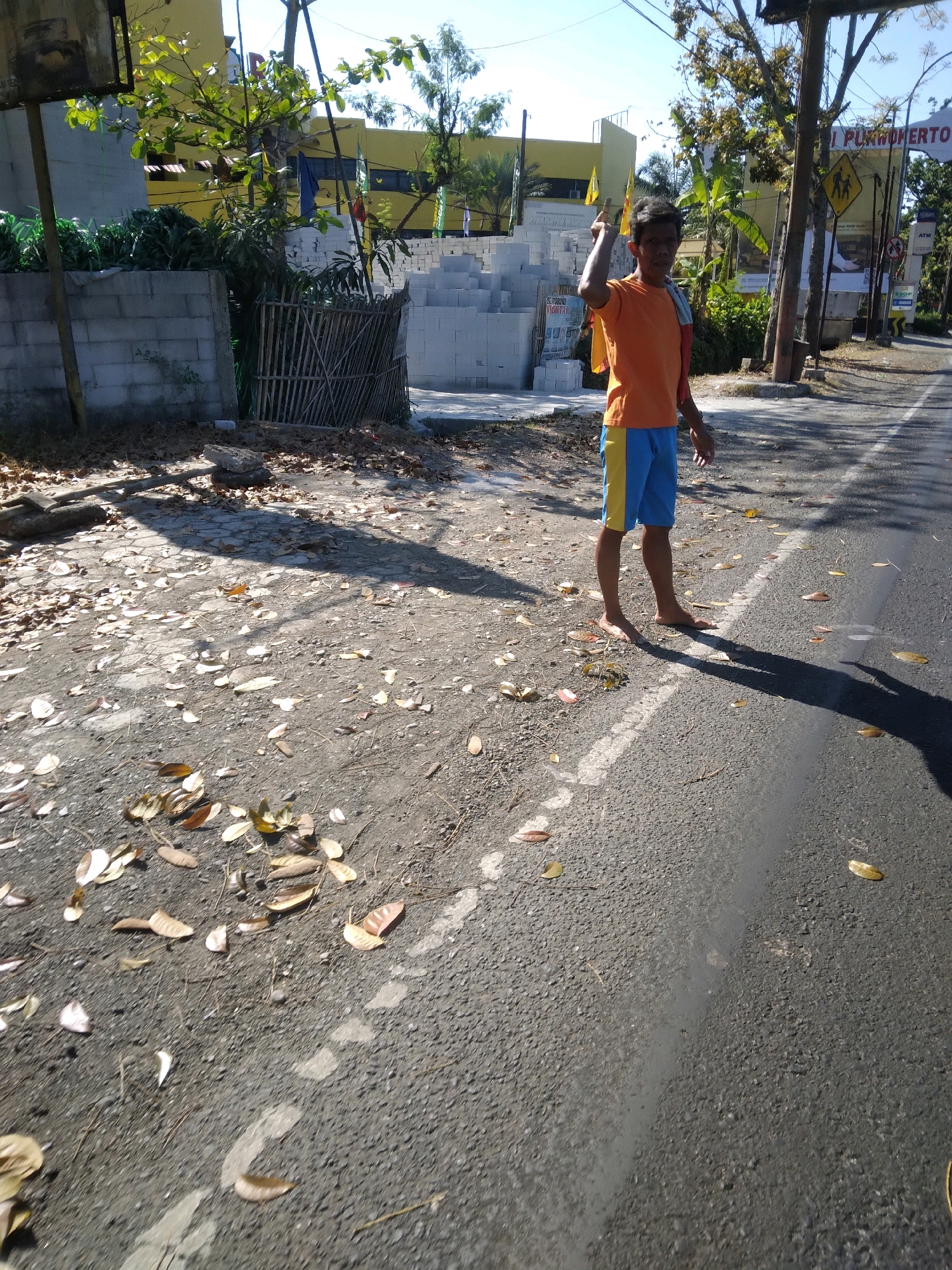 In Sokaraja, Indonesia, a young boy stands barefoot on the side of a paved road, looking towards the camera. He is wearing an orange t-shirt and blue and yellow shorts, with a yellow cloth draped over his shoulder. He is holding a piece of yellow paper in his right hand and has his left index finger raised. The road is bordered by a dirt shoulder covered in dry leaves and scattered stones. Behind the boy, a concrete wall and a bamboo fence are visible, with stacks of white cinder blocks beyond them. A yellow building with a blue roof is in the background, along with trees and utility poles. Several street signs are also present, including a yellow pedestrian crossing sign. The sun is shining, casting shadows across the scene.