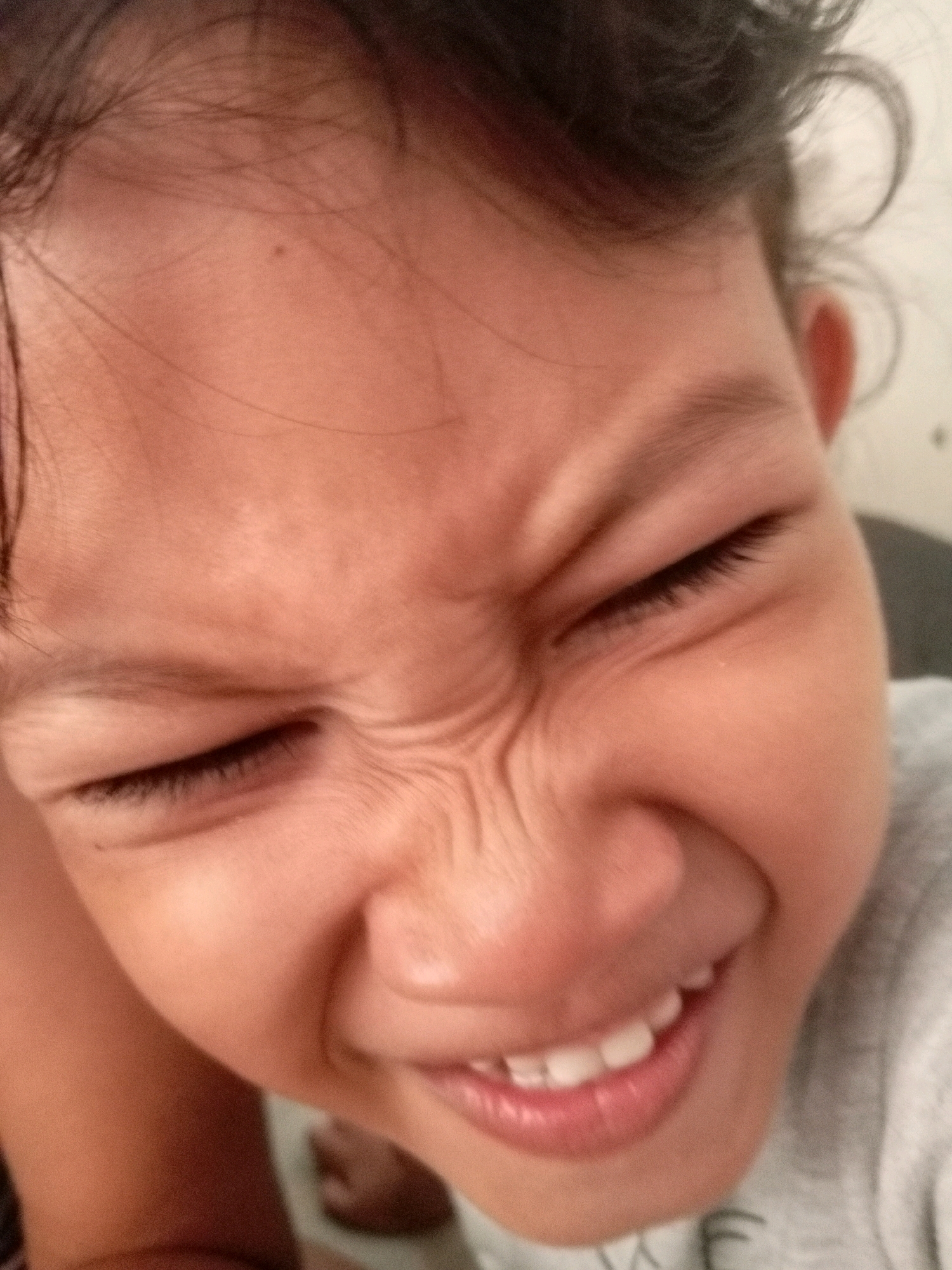 The image is a close-up shot of a young child's face, with their eyes squeezed shut and a wide grin. The child is wearing a gray shirt. The background is blurry and appears to be indoors. There is no discernible information about the location within Batam, Indonesia.
