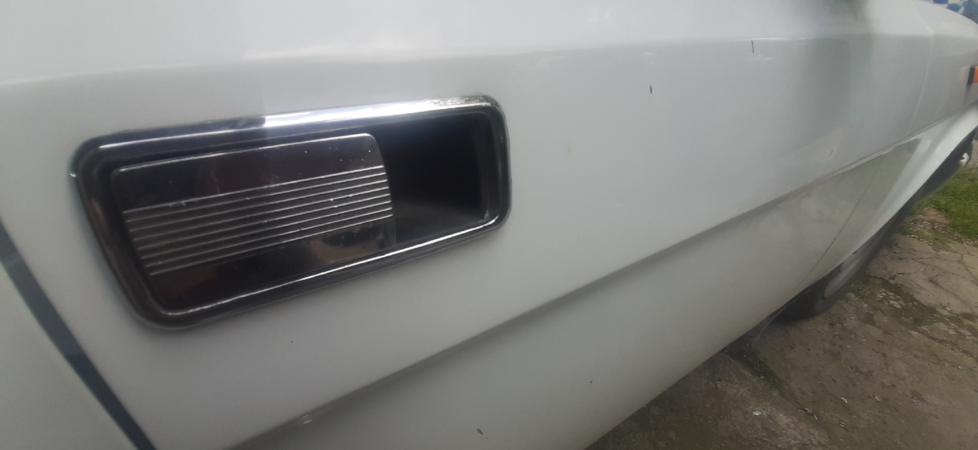 The image shows a close-up of the exterior of a white car, specifically the door handle. The door handle is chrome-plated with horizontal ridges on the inner surface. It is recessed into the car's body, which is a bright white and appears to be clean, though a few small specks are visible on the surface. The image is taken at an angle, revealing the side profile of the car. Part of a tire and the ground, which looks like a dirt or gravel path, are visible at the bottom right. The lighting suggests it is daytime, with natural light illuminating the car. The scene appears to be outdoors, possibly in a driveway or on a street in a rural or suburban setting. No people or specific events are depicted. There is no visible text in the image. The location context provided is Yessentukskaya, Russia.