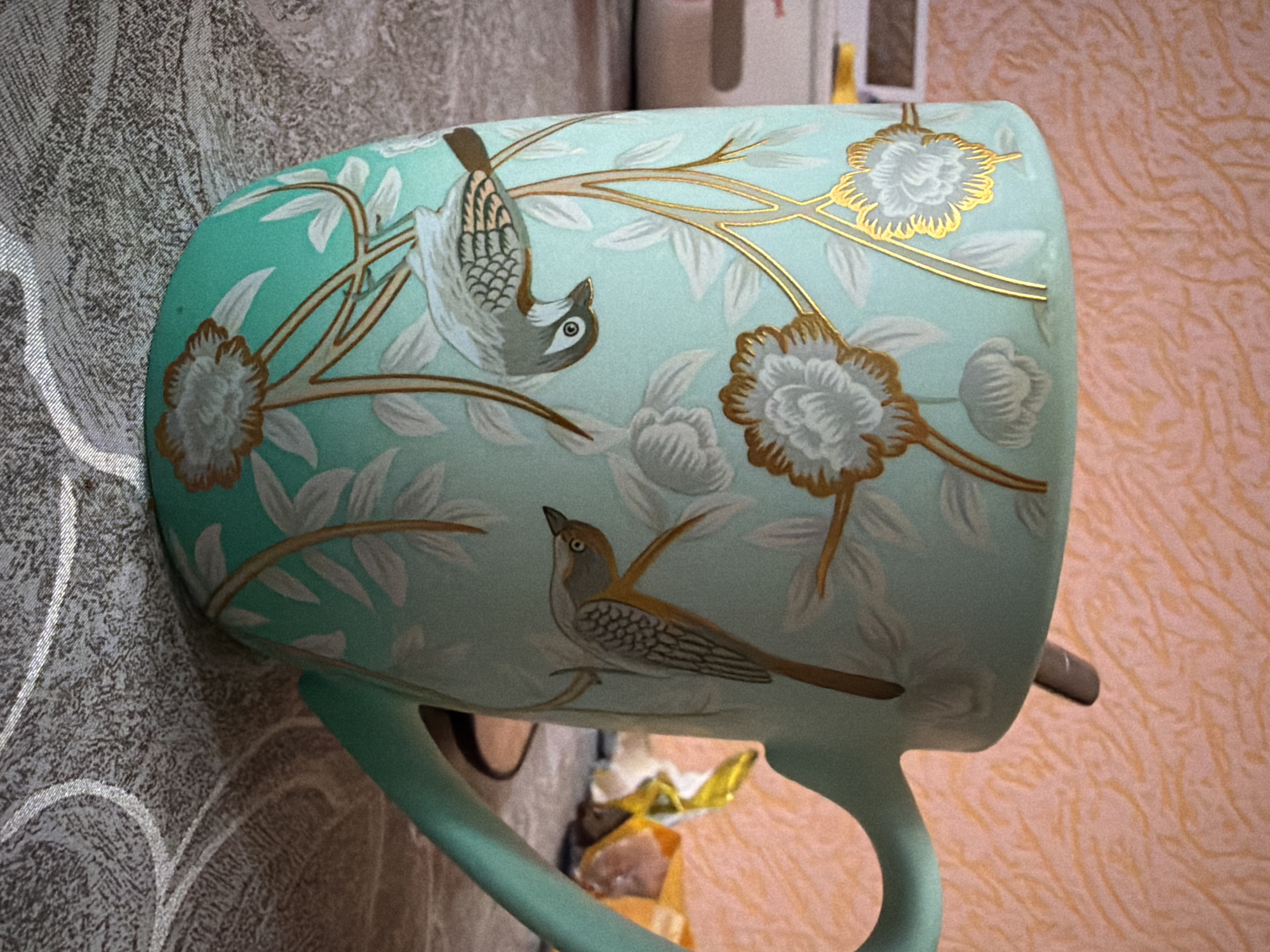 A light blue mug with a bird and flower design is depicted in the image. The mug is adorned with illustrations of two birds and several flowers, with gold accents highlighting the branches and flower details. The mug handle is also light blue. The mug is sitting on a surface with two different textures in the background: one is a wall with a swirling, peach-toned pattern, and the other is a textured surface with a gray and white swirling pattern. The lighting in the image appears to be artificial.