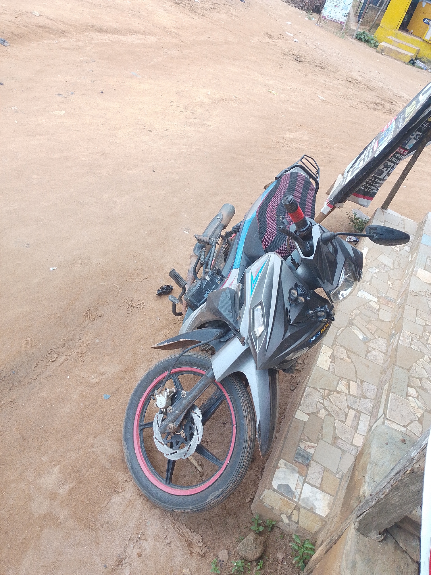 The image shows a grey and black motorcycle lying on its side on a dirt and gravel surface. The motorcycle appears to be parked or has fallen next to a tiled structure, possibly a step or a low wall. The tiled structure has a mosaic pattern made of various colored stones and tiles. In the background, there is a yellow building and a sign with text on it. The ground is sandy and appears to be unpaved. There is some vegetation growing near the tiled structure. The scene suggests an outdoor setting, possibly in a neighborhood with unpaved roads and buildings. Based on the presence of the yellow building and the general environment, the location is likely in Lagos, Nigeria.