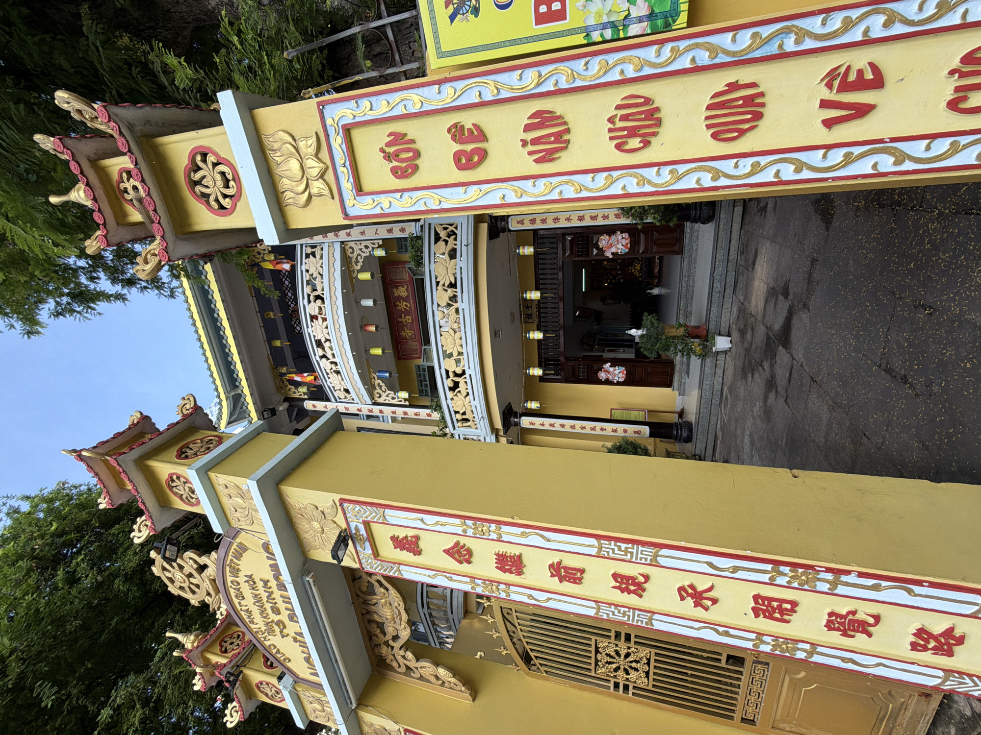 The image shows the entrance to a Buddhist temple, likely located in Nha Trang, Vietnam, given the Vietnamese script visible on the signage. The entrance is ornate and predominantly yellow, with red and gold accents. Two large yellow pillars flank the entrance, each adorned with vertical inscriptions. The pillar on the right displays the words "BON BE NAM CHAU VE" in red, raised lettering. The pillar on the left has similar red lettering in Vietnamese script.
The temple building itself is multi-tiered with yellow and green roofs, and elaborate balconies with decorative railings. Chinese or Vietnamese lanterns hang from the balconies. A large golden Dharma wheel symbol is prominently displayed above the entrance archway, along with other inscriptions in Vietnamese.
To the left of the entrance is a tall, ornate golden gate. To the right, a bright yellow sign with green and white accents is partially visible, also featuring lotus motifs and what appears to be a cartwheel symbol.
The ground in front of the temple is paved and appears slightly wet in places. Green trees and foliage surround the temple, suggesting a natural setting. No people are clearly visible within the frame of the entrance area, though it's possible people are inside the temple or just out of view. The overall impression is one of a well-maintained and visually striking religious establishment.