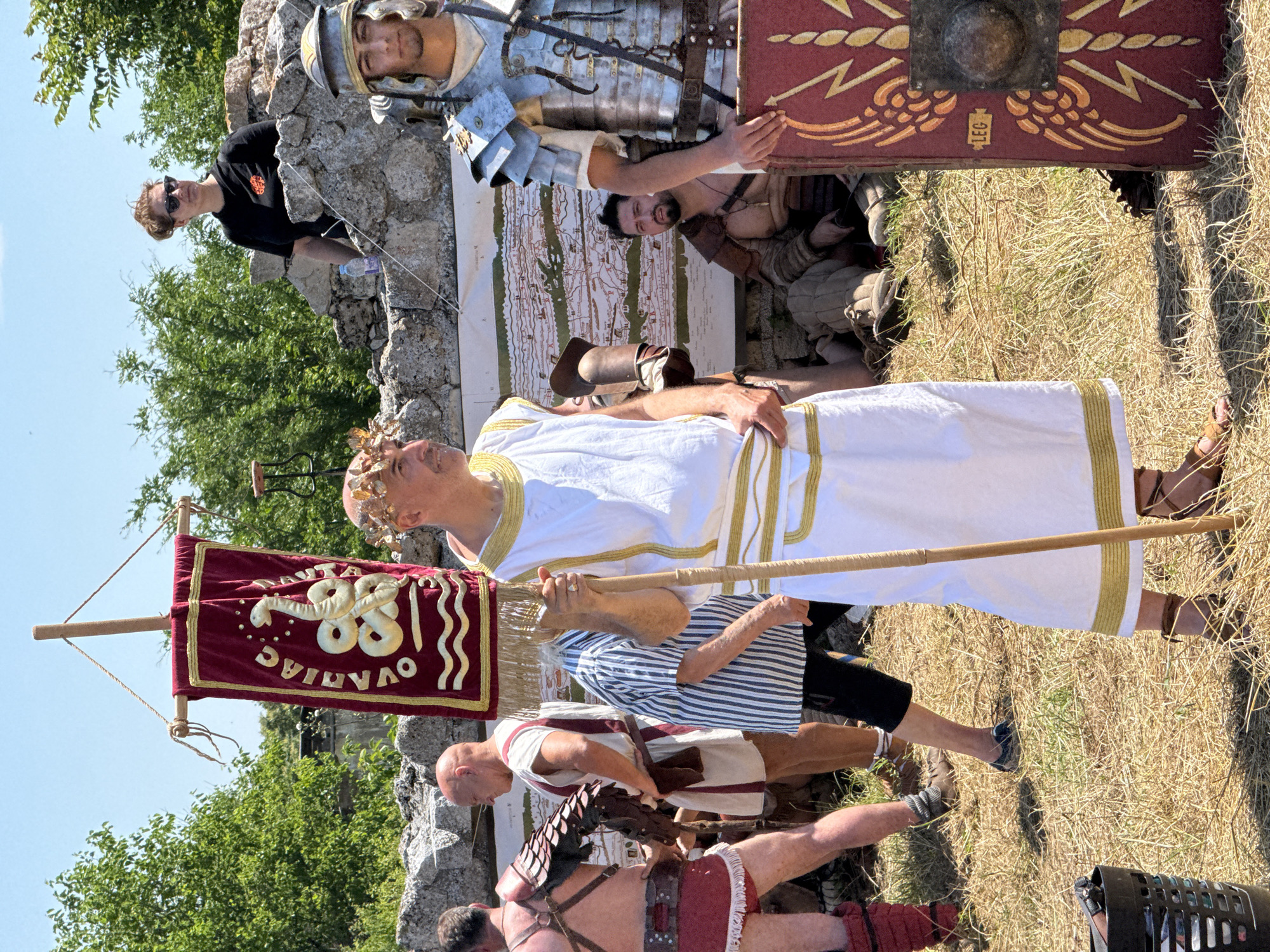 The image depicts an outdoor historical reenactment in Gigen, Bulgaria, under a clear blue sky on a sunny day. The ground is covered with dry, yellowed grass.
In the center, a light-skinned man with a balding head wears a white toga with gold trim and brown sandals, adorned with a golden leafy wreath on his head. He holds a long wooden staff from which a red rectangular banner hangs, trimmed in gold. The banner displays "OVANIAC" vertically in gold letters on the left side, a stylized gold snake resembling an ouroboros in the center, and two gold wavy lines at the bottom. The man looks towards the right.
To the right of the man in the toga, a Roman soldier stands, wearing a silver-colored lorica segmentata armor, a helmet, and a tunic. He holds a red rectangular shield marked "LEG" on a small yellow plaque, adorned with gold lightning bolt and wing patterns, and a central metal boss. The soldier looks forward.
To the left of the man in the toga, a man wearing a vertical blue and white striped shirt and dark pants is partially visible, bending forward. Further left, another man in red leg coverings and a partial brown armor piece is visible from the waist down. Other men in simple white tunics and brown footwear are present in the midground.
In the midground, a low stone wall or ruin extends horizontally. Behind this wall, a bearded man with dark hair is seated on the ground, wearing brown leather armor and a necklace, looking left. A large map is displayed on a white board behind him.
On top of the stone ruins in the background, a woman with light brown hair, wearing a black t-shirt with a red emblem and sunglasses, observes the scene, looking towards the right. Green trees are visible in the far background beneath the bright sky. A black mesh basket is partially visible in the bottom-left corner of the frame.