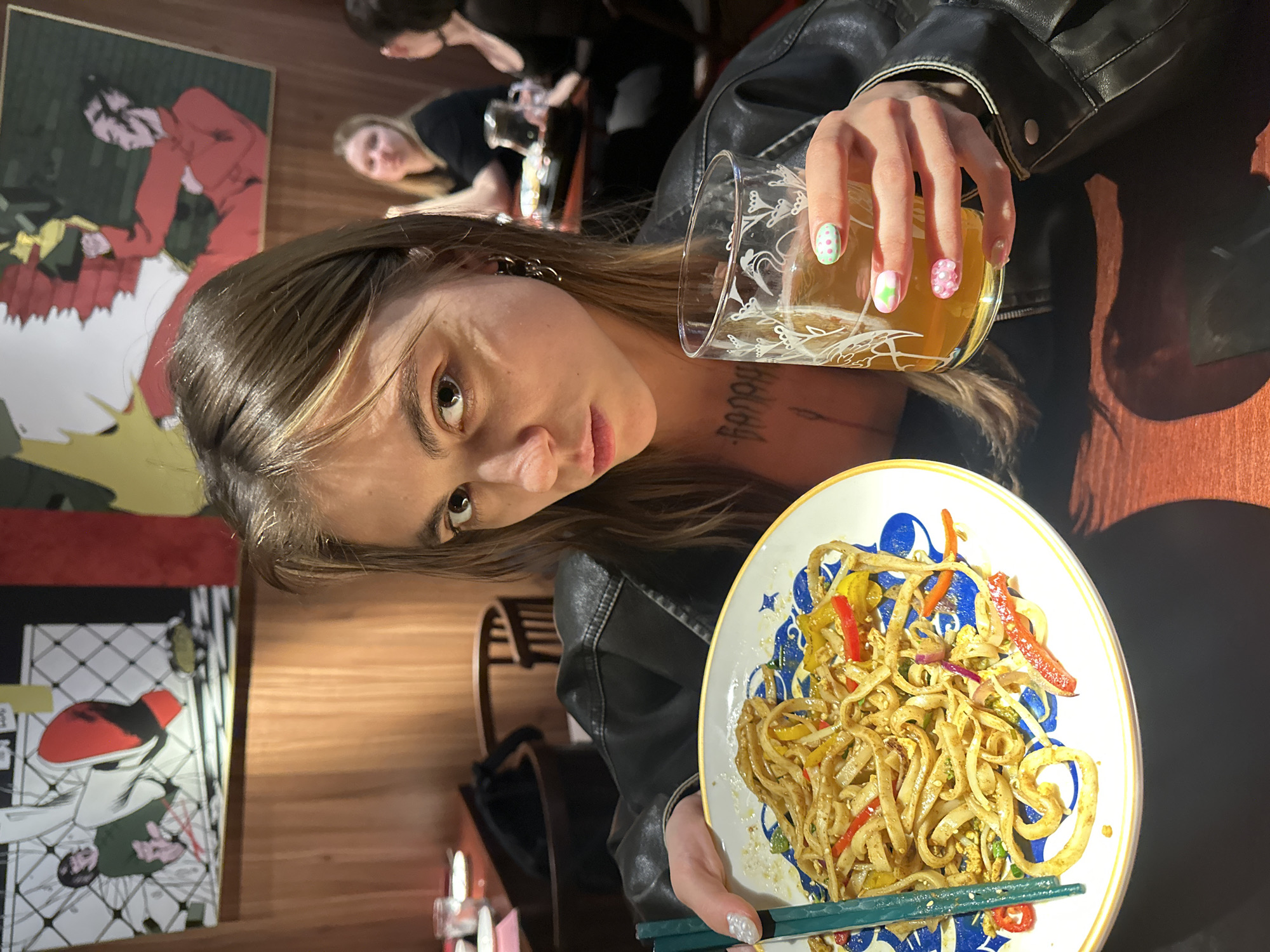 A young woman with shoulder-length brown hair and a black leather jacket is sitting at a table in a restaurant, holding a plate of noodles and a glass of beer. She is looking directly at the camera with a neutral expression. The restaurant has wooden paneling on the walls and artwork depicting cartoon-like figures in comic book style. In the background, other patrons can be seen at tables. The overall setting appears to be a casual dining establishment. The image does not provide any information to suggest it is located in Moscow, Russia.