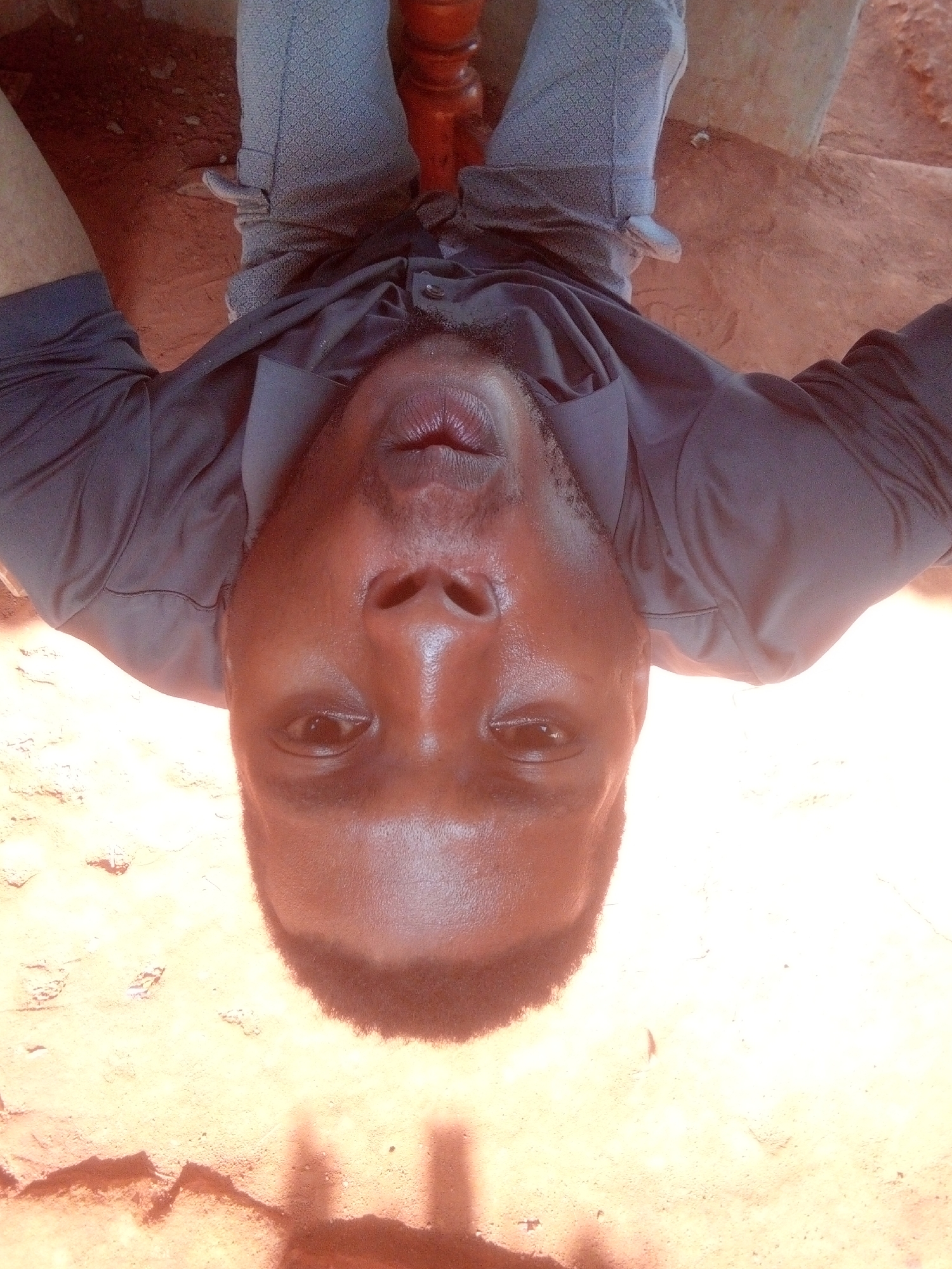 A close-up, upside-down perspective photograph shows a dark-skinned man wearing a dark, collared shirt and light-colored patterned pants. He is positioned on or near a wooden object, possibly a chair. The background is a light, sandy color. The image appears to have been taken outdoors, with some areas overexposed. The location is Unknown, Unknown.