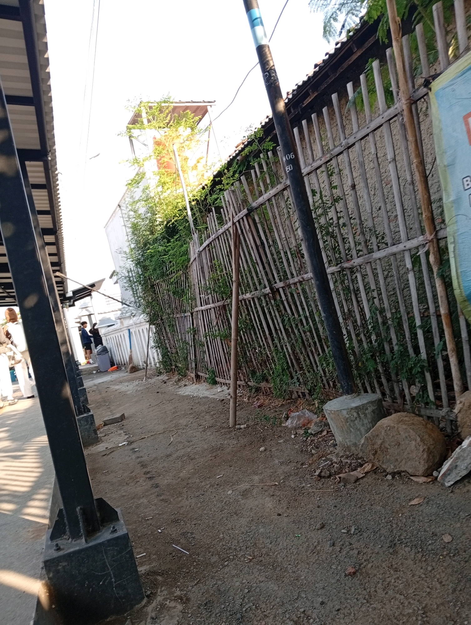 The image depicts an outdoor scene, likely in an urban or semi-urban area of Karawang, Indonesia. The foreground is dominated by a dirt and gravel path leading towards a bamboo fence that runs along the right side of the frame. A dark metal pole, possibly a lamppost, stands in the middle ground, its base obscured by debris. Behind the fence, lush green vegetation grows. To the left, a covered walkway with a corrugated metal roof is visible, supported by dark metal pillars. In the distance, a white building with a rusty metal structure on top is partially obscured by trees. Several people are present: two individuals stand near a trash can and a broom leaning against a white structure on the left side of the path, while a third person, wearing a hijab, walks away from the camera on the far left. The lighting suggests it is daytime, with shadows cast by the walkway structure. The overall impression is one of a functional outdoor space, possibly near a residential or commercial area, with elements of both infrastructure and natural growth.
