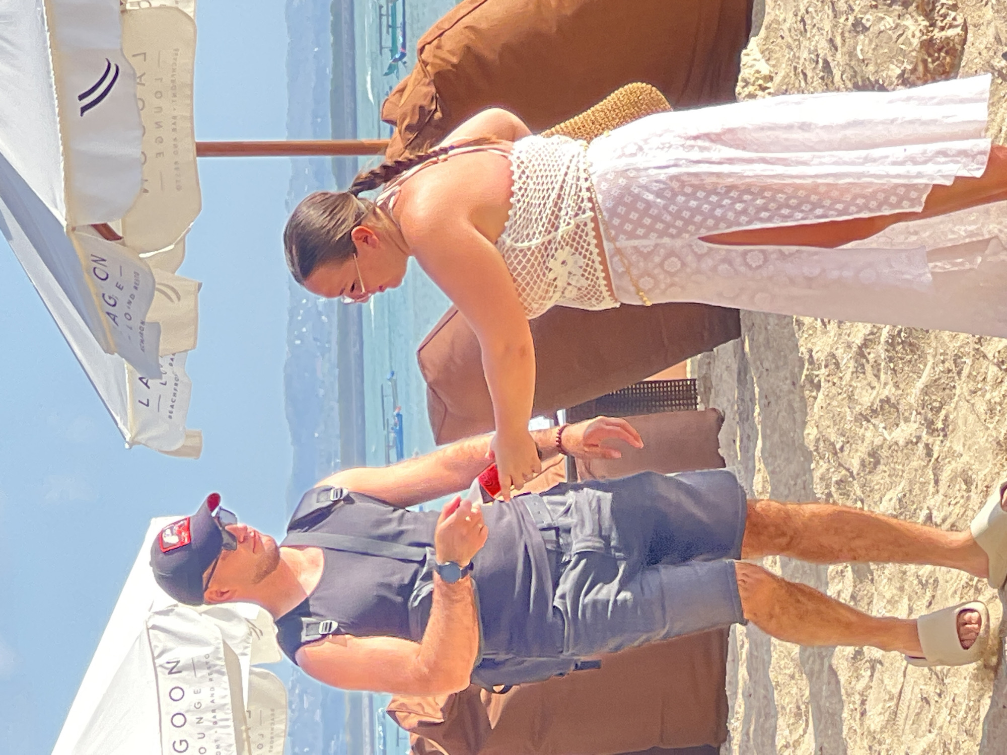 The image shows a man and a woman standing on a sandy beach in Kuta, Indonesia. The man, wearing a black tank top, grey shorts, and a baseball cap, is holding a small red object, possibly a ring box, which the woman, in a white crocheted top and a white skirt with a thigh-high slit, is reaching for. They are positioned between beach umbrellas and large brown beanbag chairs, with the ocean and distant hills visible in the background. The scene suggests a romantic moment on a sunny day at a beachside establishment in Kuta.