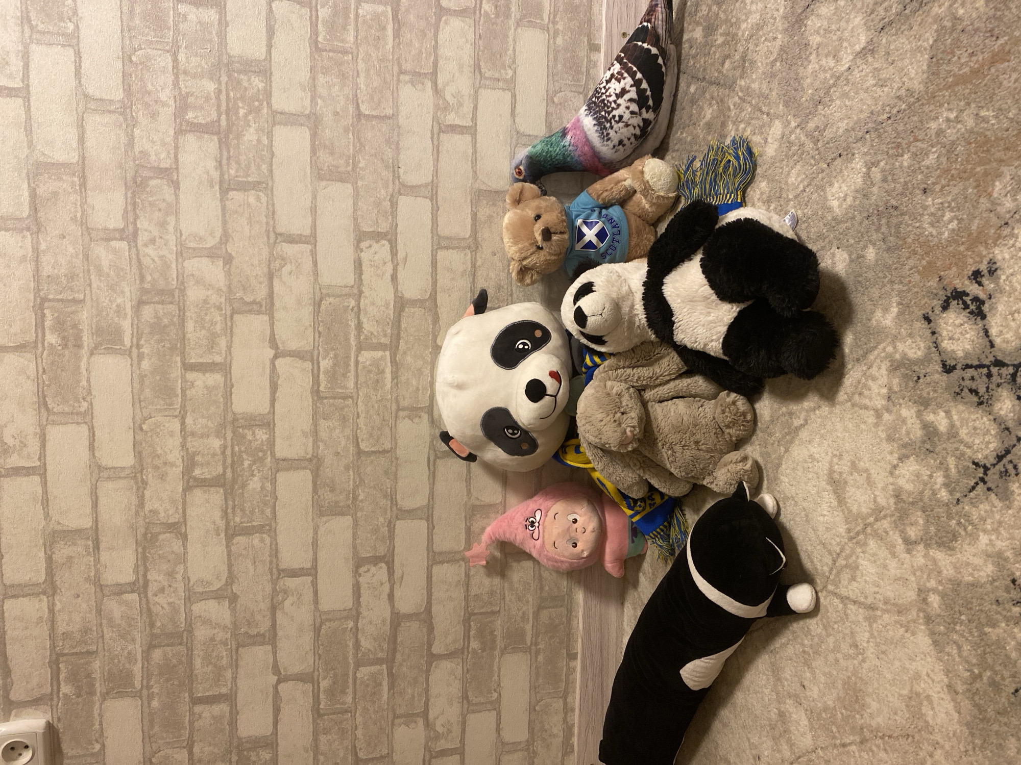 A group of stuffed animals and a taxidermied bird are arranged on a patterned rug in front of a brick-patterned wallpaper. From left to right, a long black and white stuffed cat lies on the floor. Behind it, a small pink stuffed doll with a pointed hat is propped up. To its right is a large stuffed panda, wearing a blue and yellow striped scarf. In front of the panda, a plush bunny sits. To the right of the bunny and slightly behind it, a small brown teddy bear wears a blue shirt with a white saltire. next to the teddy bear, a taxidermied pigeon is positioned with its head angled towards the bear. A smaller stuffed panda is in the foreground, facing away from the camera. The lighting appears to be artificial, casting soft shadows on the subjects and the rug.
