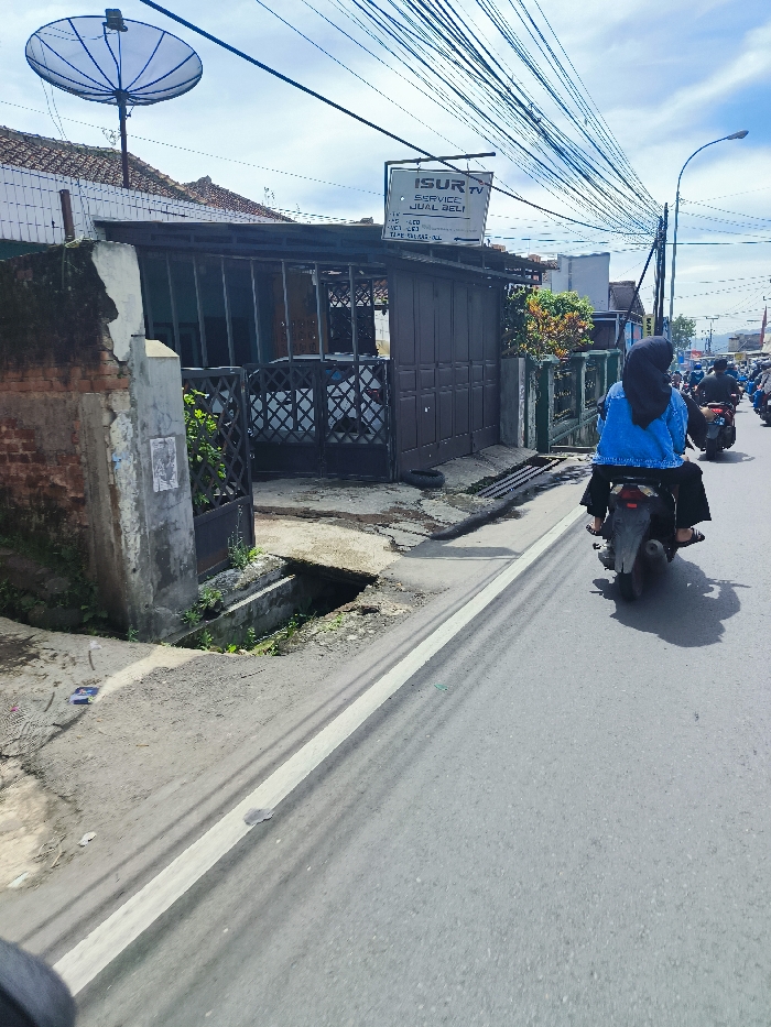 The image shows a street in Ciwidey, Indonesia (latitude: -7.104, longitude: 107.46), with a person riding a motorcycle on the right side of the road. The road is paved and has a white line separating it from the sidewalk. A small, partially visible car is parked in the driveway of a small, single-story brick house. The motorcycle is in the middle of the road. There are multiple power lines strung across the scene. The person on the motorcycle is wearing a blue shirt and a black hijab and cannot be seen from the waist down. There is a partially visible sign that says "ISUR TV Service Jual Beli". The street is empty except for the motorcycle. There is a small drainage ditch along the curb. The weather is clear and sunny.