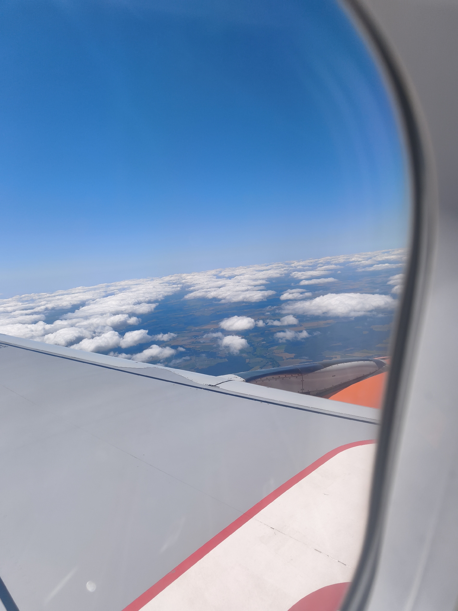 An aerial view from an airplane window looks out over a landscape of clouds, fields, and some wind turbines under a clear blue sky. The left side of the frame is dominated by the gray wing of the airplane, with a red and white stripe visible near the bottom. The right side of the frame shows the edge of the airplane window. The scene is a typical view from a commercial flight, offering a glimpse of the world from above. There is no indication of the city of Golßen, Germany, in this image.
