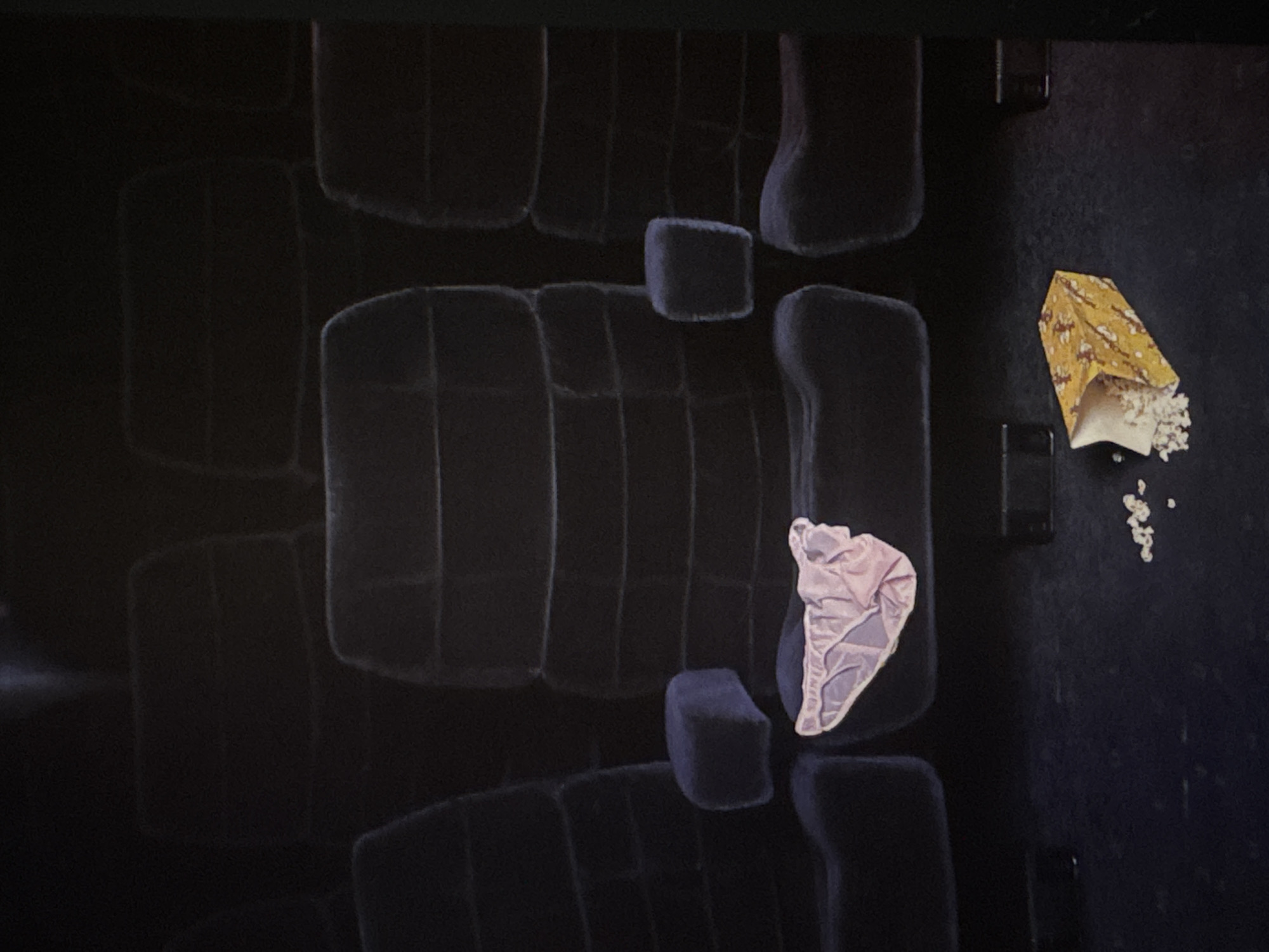 The image shows a row of empty, dark blue theater seats. On one of the seats in the foreground, a pair of pink lingerie is placed. In front of the seats, on the floor, is an overturned box of popcorn with some kernels spilled out. The scene is dimly lit, typical of a movie theater. There is no visible indication of the location in Kuta Selatan, Indonesia.