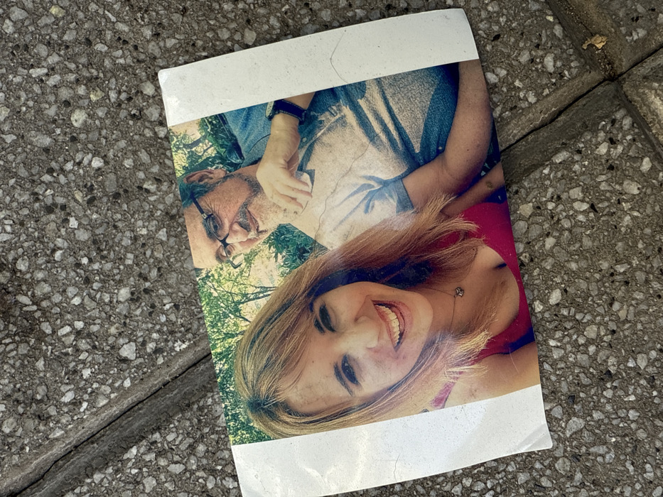 A discarded photograph lies on a textured stone surface. The image depicts a smiling woman in a red top, closely positioned with a man wearing glasses and a casual shirt. Greenery suggests an outdoor setting. The photo has a thick white border and appears slightly worn, with visible imperfections. The orientation is tilted, adding to the sense of abandonment.