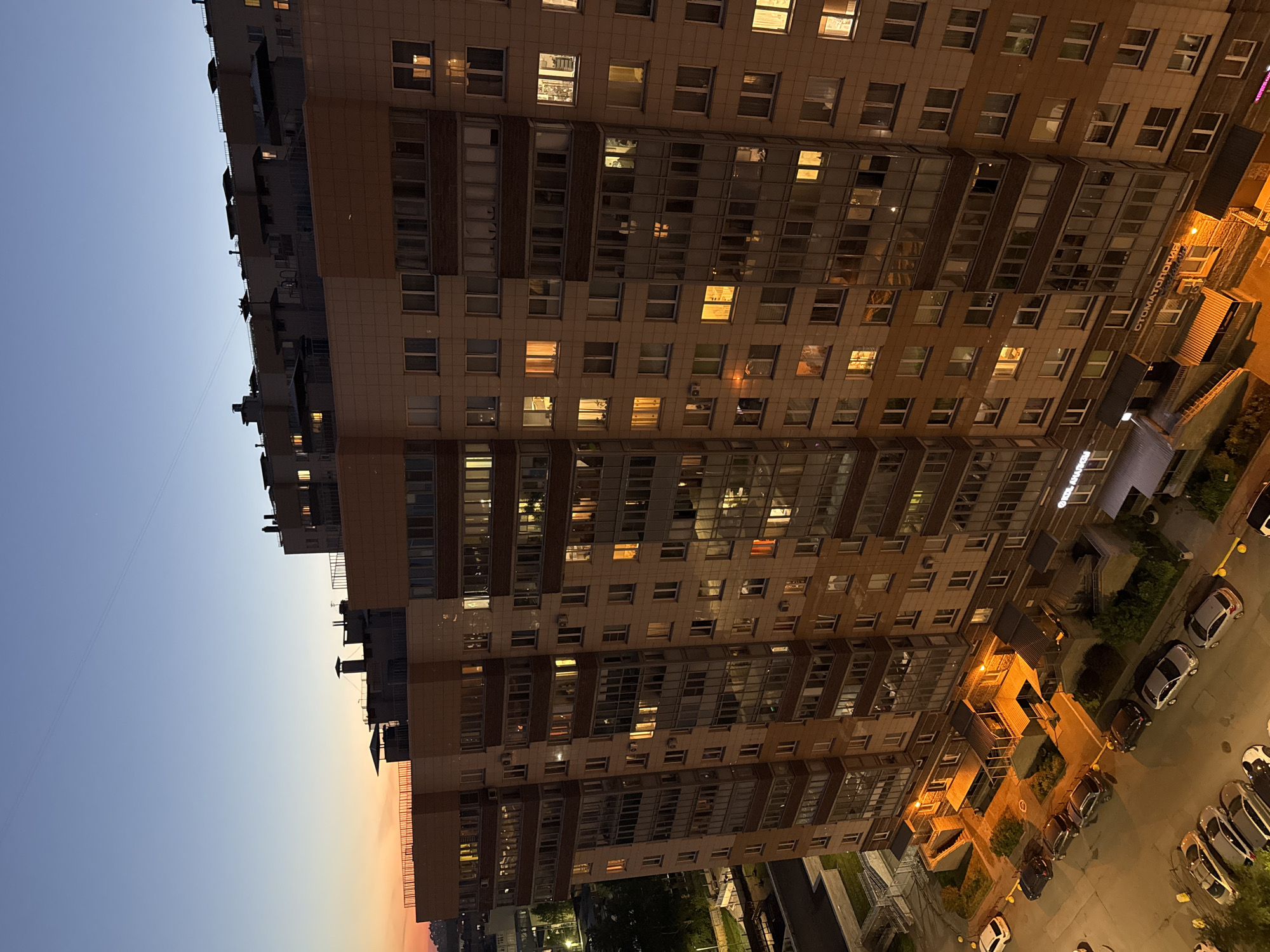 The image captures a tall, modern residential or mixed-use building at twilight or dawn, viewed from a high vantage point looking down its facade and the street below.
The building is constructed with light-toned panels, possibly concrete or brick, featuring numerous rectangular windows. Many windows across various floors are illuminated with warm, yellow light, suggesting active interiors. Several floors incorporate extensive glass-paned balconies or loggias, which reflect the indoor lighting.
At ground level, a street or parking area is visible with several parked cars. Streetlights emit bright orange light, illuminating the ground and vehicles. Commercial establishments occupy the lower floors of the building. A prominent sign on one of these units clearly displays "СТОМАТОЛОГИЯ" (Stomatologiya), indicating a dental clinic. Another partially visible sign on an adjacent or distant structure appears to read "ОТЕЛЬ АПАРТАМЕНТЫ" (Otel Apartamenty), suggesting a hotel or serviced apartments.
The sky, occupying the left side of the frame, shows a gradient of colors characteristic of twilight: a deep blue at the top transitioning to a lighter, pale orange-pink hue near the horizon, indicative of either sunset or sunrise. The weather appears clear with no clouds. A faint thin line, possibly a wire, is visible against the sky. The scene is quiet and calm, typical of an urban setting during these hours in Russia.