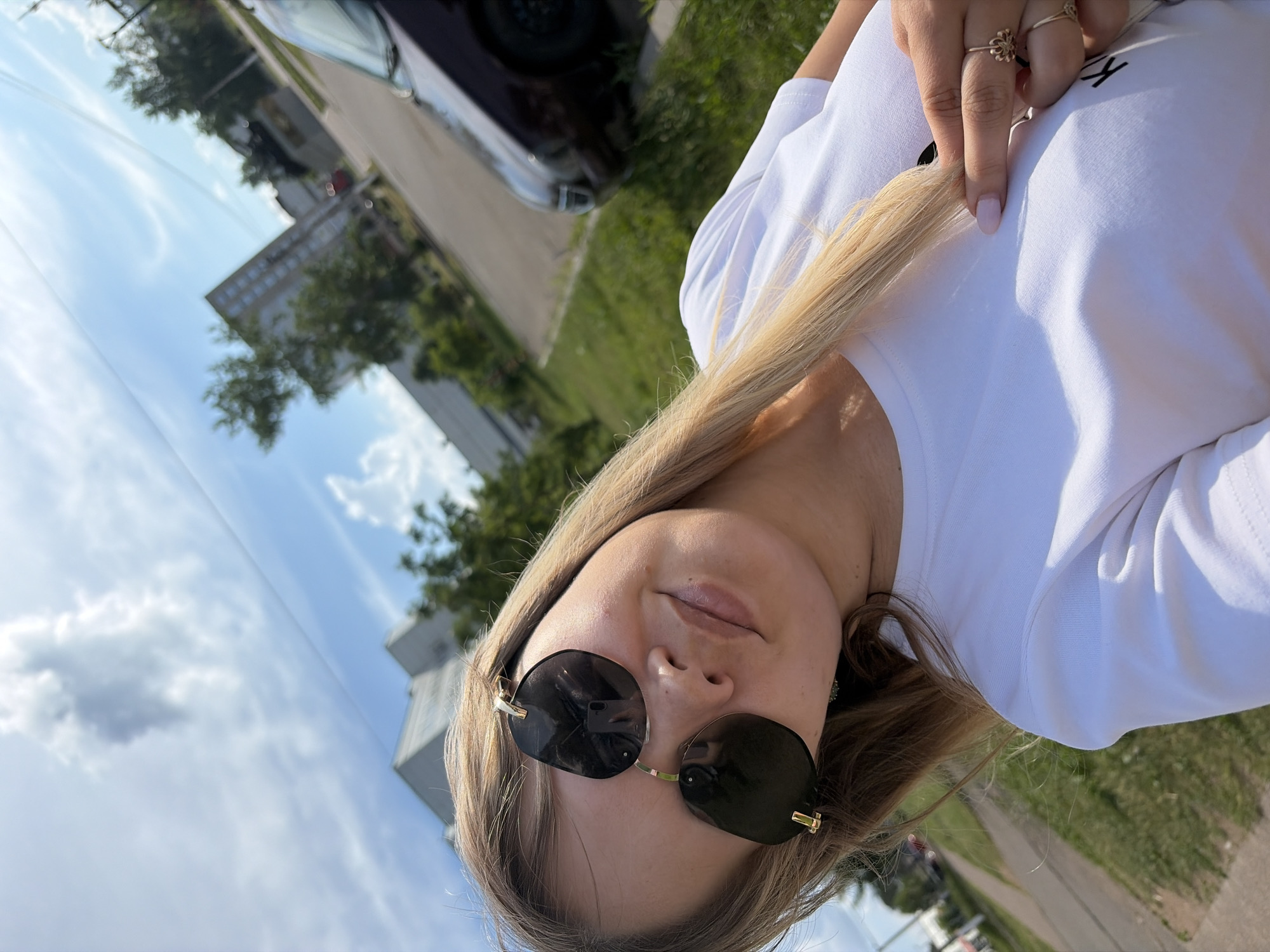 A young woman with blonde hair and sunglasses is taking a selfie outdoors in the city of Chaikovsky, Russia. She is wearing a white t-shirt. Her right hand is visible, with rings on her fingers, and she is touching her hair. In the background, there are buildings, trees, a road, a parked car, and a sidewalk with a grassy area. The sky is blue with some clouds.