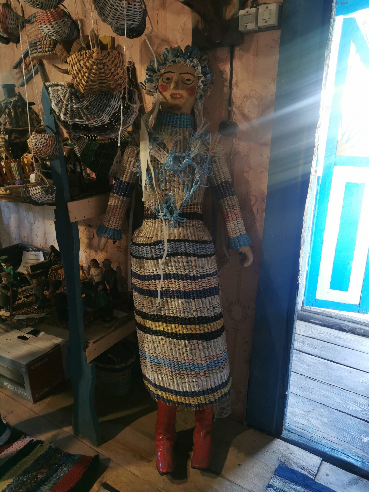 The image depicts a full-body, upright, woven effigy resembling a person, adorned with a blue and white headdress, blue beaded necklace, and a striped woven dress in shades of blue, white, yellow, and black. The effigy wears red boots and stands in a room with a light-colored, patterned wallpaper. To the left of the effigy, shelves are visible, holding various woven baskets and what appear to be figurines. A dark blue wooden support structure is also present. To the right, a bright blue-framed window is partially open, allowing sunlight to stream into the room and cast a glare. The floor is made of wooden planks. The context suggests this scene is located within a building in the Шчарчоўскі сельскі Савет, Belarus, likely a shop or a cultural display area showcasing traditional crafts.