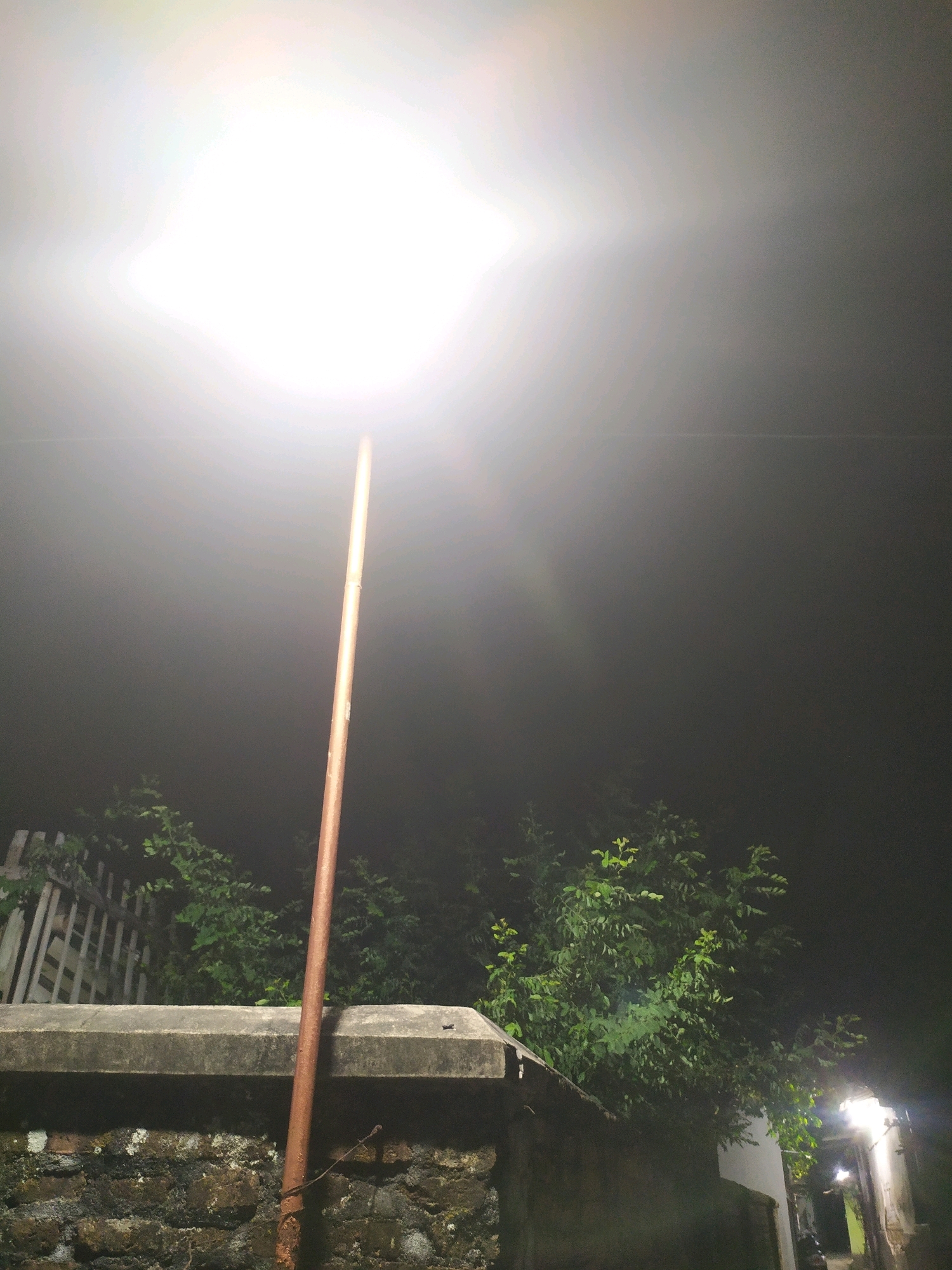 The image depicts an outdoor scene at night, likely in a residential area. A tall, thin lamppost with a rusted metal pole stands vertically in the foreground, illuminated by its own bright light source at the top, which is the dominant light in the frame. The light creates a strong lens flare and washes out the upper portion of the image. Below the lamppost, a textured wall, constructed from rough stones and mortar, forms the lower part of the scene. Above this wall, lush green foliage from trees is visible, extending upwards and to the right. A white wooden fence is partially visible on the left, behind the foliage. In the background, a pathway is illuminated by a second, smaller light fixture, suggesting it leads to a building or another part of the property. The overall atmosphere is dim and hazy, possibly due to mist or fog, which softens the details of the background. The setting appears to be Glagahwangi, Indonesia, based on the provided context.