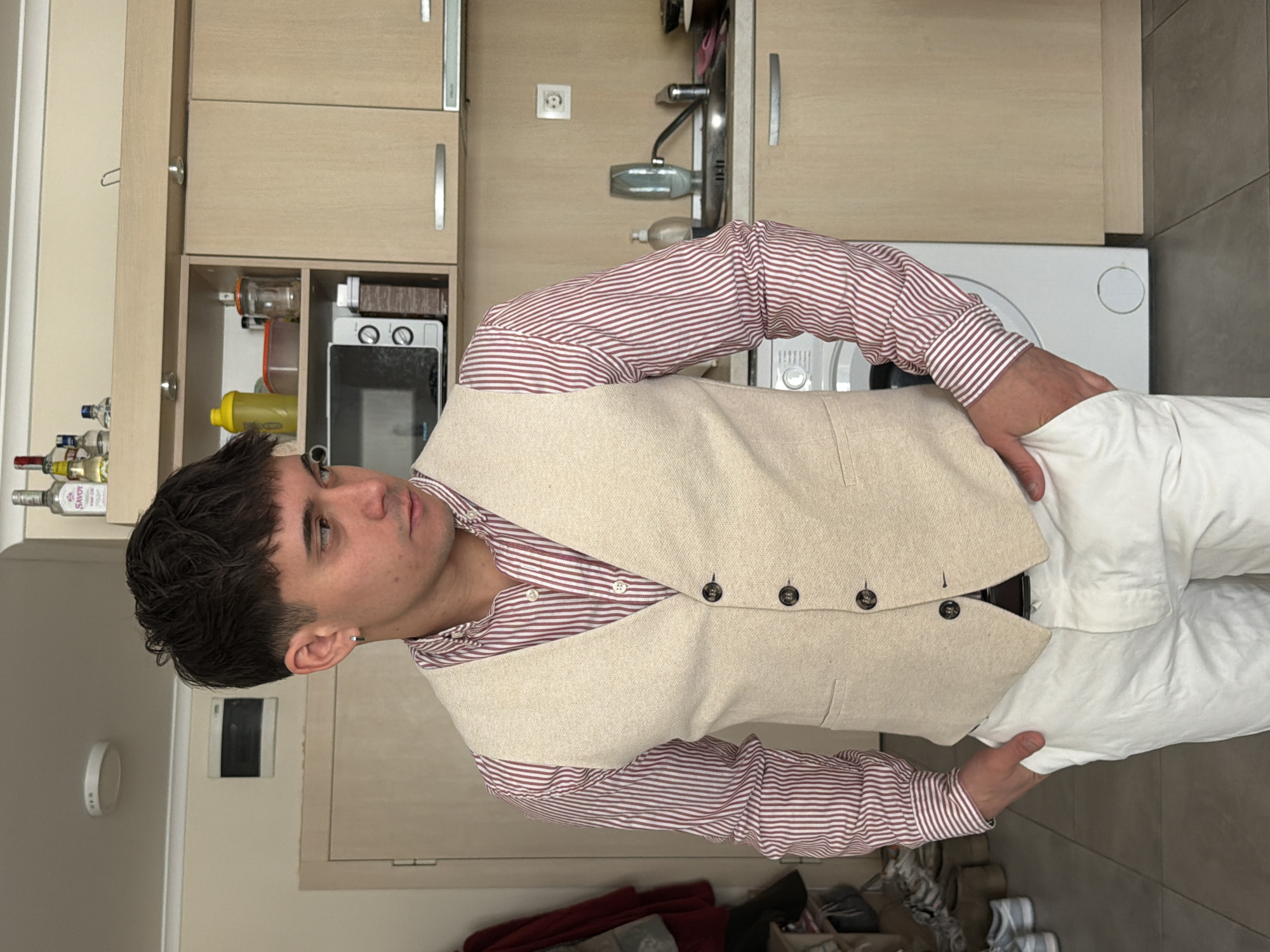 A young man stands in a room that appears to be a kitchen in Sofia, Bulgaria. He is wearing a striped red and white long-sleeved shirt, a beige waistcoat, and white trousers. He is partially facing the camera with his head slightly tilted. His left hand is in his trouser pocket, while his right hand is holding the fabric of his trousers. Behind him is a kitchen counter and cabinets in a light wood color. A microwave is visible, as well as various bottles of liquids on a shelf. To his right, there is a white appliance, possibly a washing machine or dryer. The floor is tiled. Some shoes and other items are visible near the bottom of the image. The lighting appears to be soft and even, suggesting natural light from a window. A light switch or sensor is visible on the wall to the left of the subject.
