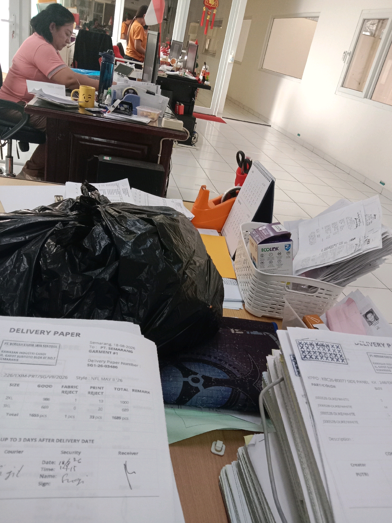 The image depicts an office environment at PT. Berkah Karya Jaya Sentosa, located in Kawasan Industri Candi, Jl. Gatot Subroto Blok BF No.7, Semarang, Indonesia. The foreground is dominated by a large black trash bag overflowing with what appears to be discarded papers and documents. To the left, a woman wearing a pink shirt and brown pants is seated at a dark wood desk, looking down at documents and papers. A yellow mug with a smiley face is placed on the desk. The desk itself is cluttered with various office supplies, including papers, pens, a blue water bottle, and a tape dispenser. In the midground, a white woven basket holds a stack of papers and two boxes labeled "ECOLINK". A desk calendar showing August 2026 is also visible. In the background, another person wearing an orange shirt is visible near a computer monitor. The overall impression is one of a busy, somewhat disorganized office setting.