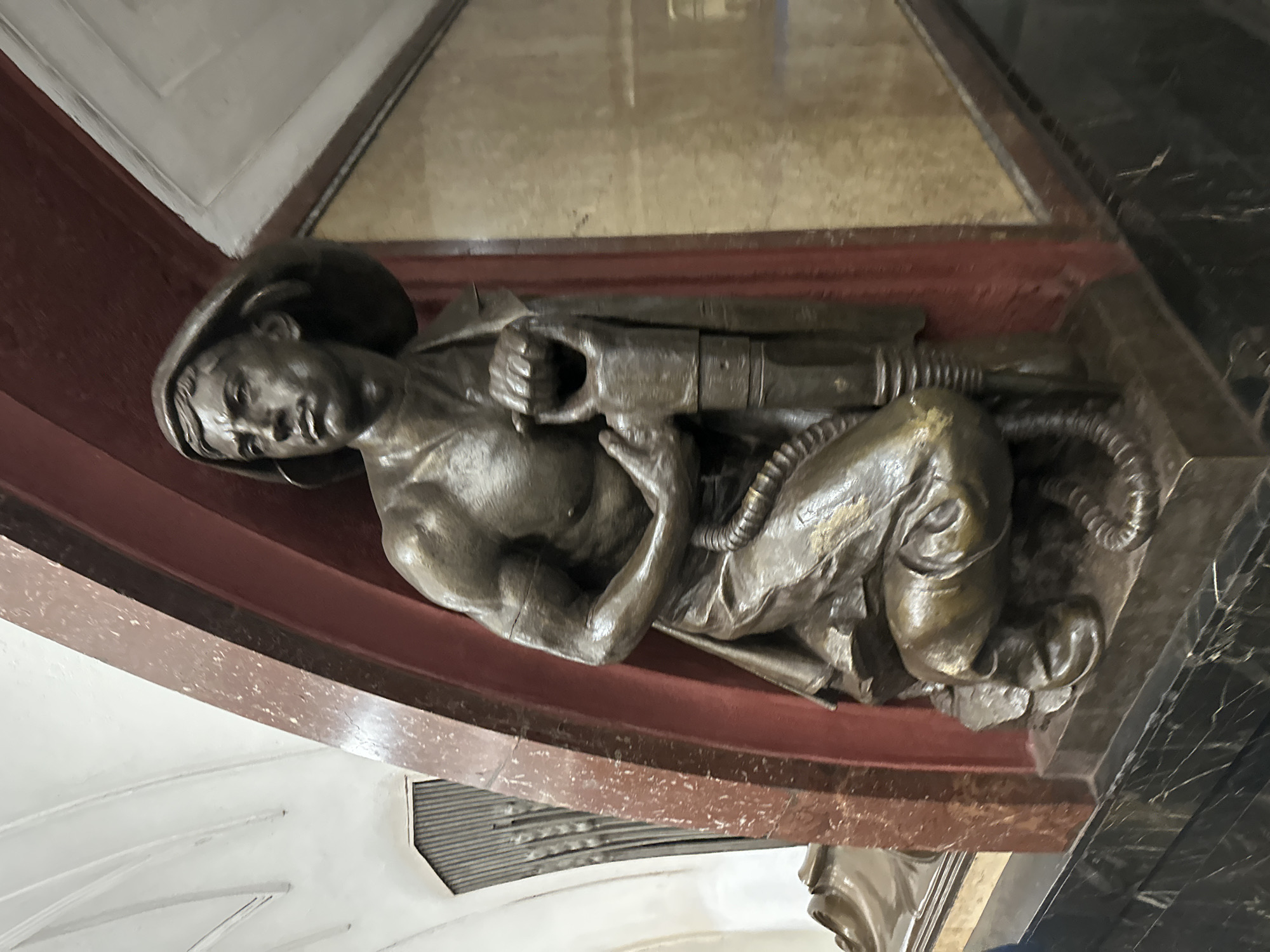 The image shows a bronze sculpture of a man wearing a hat and holding a jackhammer. The sculpture is positioned within an alcove decorated with red and beige marble. The man is kneeling and looking upwards and to the left with a determined expression. His muscular physique is emphasized by the rendering of his arms and chest. A flexible hose snakes from the jackhammer and coils around the figure. The background includes an ornate ceiling with white curved beams and a metallic grate on the left. The sculpture is mounted on a black marble pedestal. This artwork is located in the Mayakovskaya metro station in Moscow, Russia, a station renowned for its art deco style and mosaics.