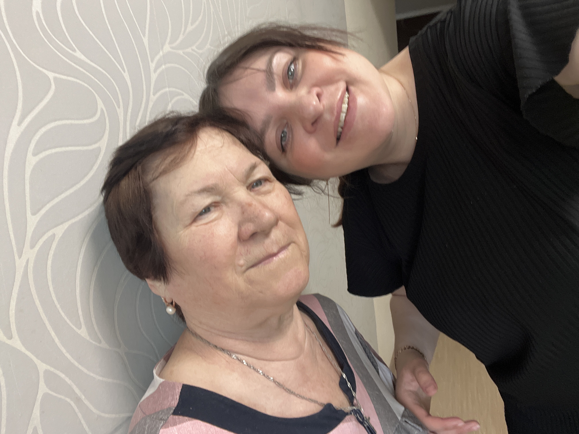 Two women are captured in a close-up, informal portrait, likely a selfie, in an indoor setting in Perm, Russia. The image features an older woman in the foreground and a younger woman positioned behind and slightly above her, leaning her head affectionately against the older woman's.
The older woman, with short, dark brown hair, smiles softly. She wears a multi-striped top with pink, black, and grey sections, and a silver chain necklace. A white pearl earring is visible in her right ear. Her face shows natural signs of aging.
The younger woman, also with dark brown hair, smiles broadly, revealing her teeth. She wears a black, ribbed top and a delicate gold chain bracelet on her left wrist. Her arm and hand are partially visible in the lower right portion of the image.
Both women appear content and happy. The background consists of a light grey wall with a subtle, stylized white pattern resembling organic shapes or leaves, suggesting a domestic or personal environment. There is no visible text in the image.