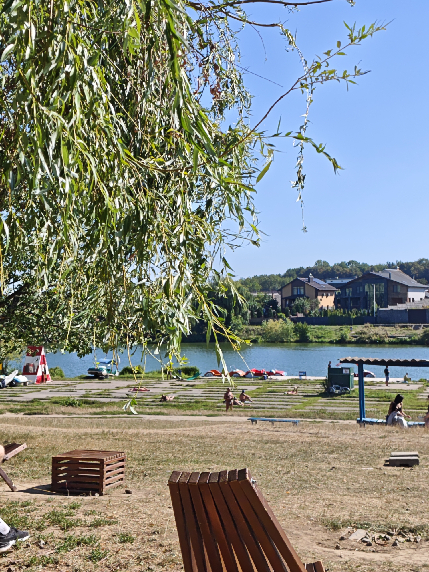 The image depicts a sunny day at a waterfront recreation area, likely in Vinnytsia, Ukraine, evidenced by the architecture of the houses in the background. The scene is dominated by a large weeping willow tree in the foreground, its long, drooping branches obscuring a portion of the view. Beyond the tree, a calm blue lake stretches across the middle ground. On the opposite shore, several houses with modern architectural styles, including some with solar panels on the roofs, are visible.
The immediate foreground and middle ground show a grassy area with a few scattered wooden benches and a small wooden crate. In the middle ground, closer to the lake, there are several colorful paddle boats lined up on the shore. A red and white striped tent-like structure and some white watercraft are also present near the water's edge.
A few individuals are visible in the scene. On the grassy area near the lake, there are people sunbathing and sitting. One person is seen walking in the foreground, wearing black shoes. Further along the shore, under a blue-roofed canopy, two individuals are standing, one appearing to be a woman in light-colored clothing. The overall atmosphere is one of leisure and outdoor activity.