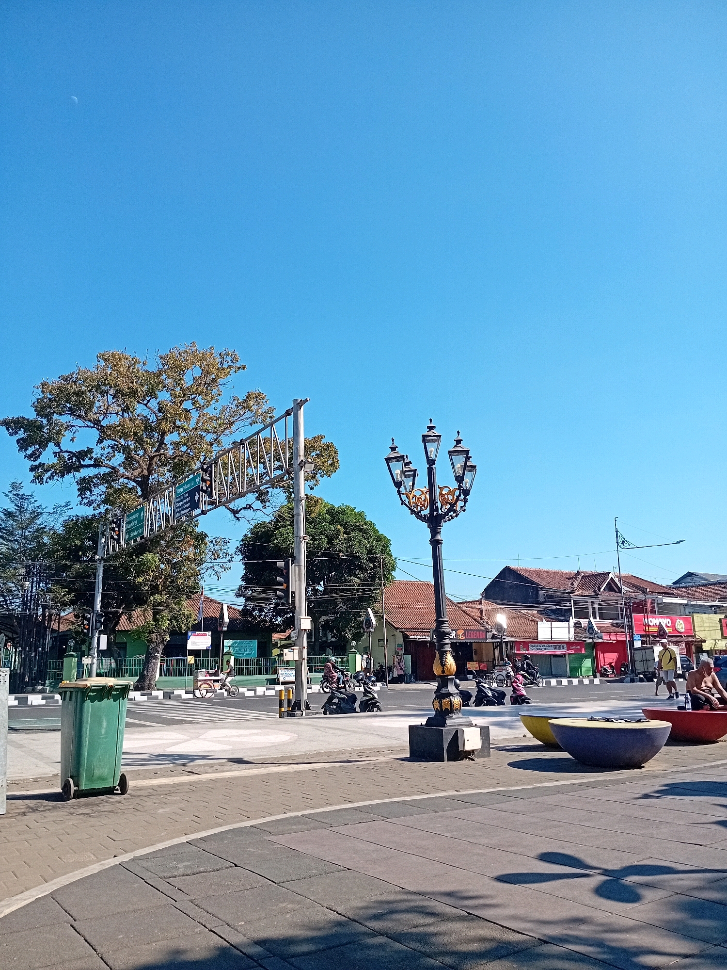 The image captures a street scene in Purworejo, Indonesia, on a clear, sunny day. In the foreground, a gray paved walkway transitions to a patterned gray and beige stone surface. To the left, a green trash bin with wheels sits on the paved area. Dominating the center is an ornate black lamppost with gold accents and multiple light fixtures. In the middle ground, a street intersection is visible with traffic lights and directional signs. A large tree with sparse, autumn-colored foliage stands prominently to the left of the signs. A row of buildings with red-tiled roofs lines the right side of the street, featuring various shops and businesses with colorful signage, including "OPWO" and "LEGENDA." Several motorcycles are parked and in motion on the street. To the far right, two large, colorful, bowl-shaped seating structures are placed, one red and one yellow, with a third purple one partially visible. A partially clothed man is seated on the red structure. The sky is a vivid, cloudless blue with a sliver of the moon visible in the upper left quadrant.