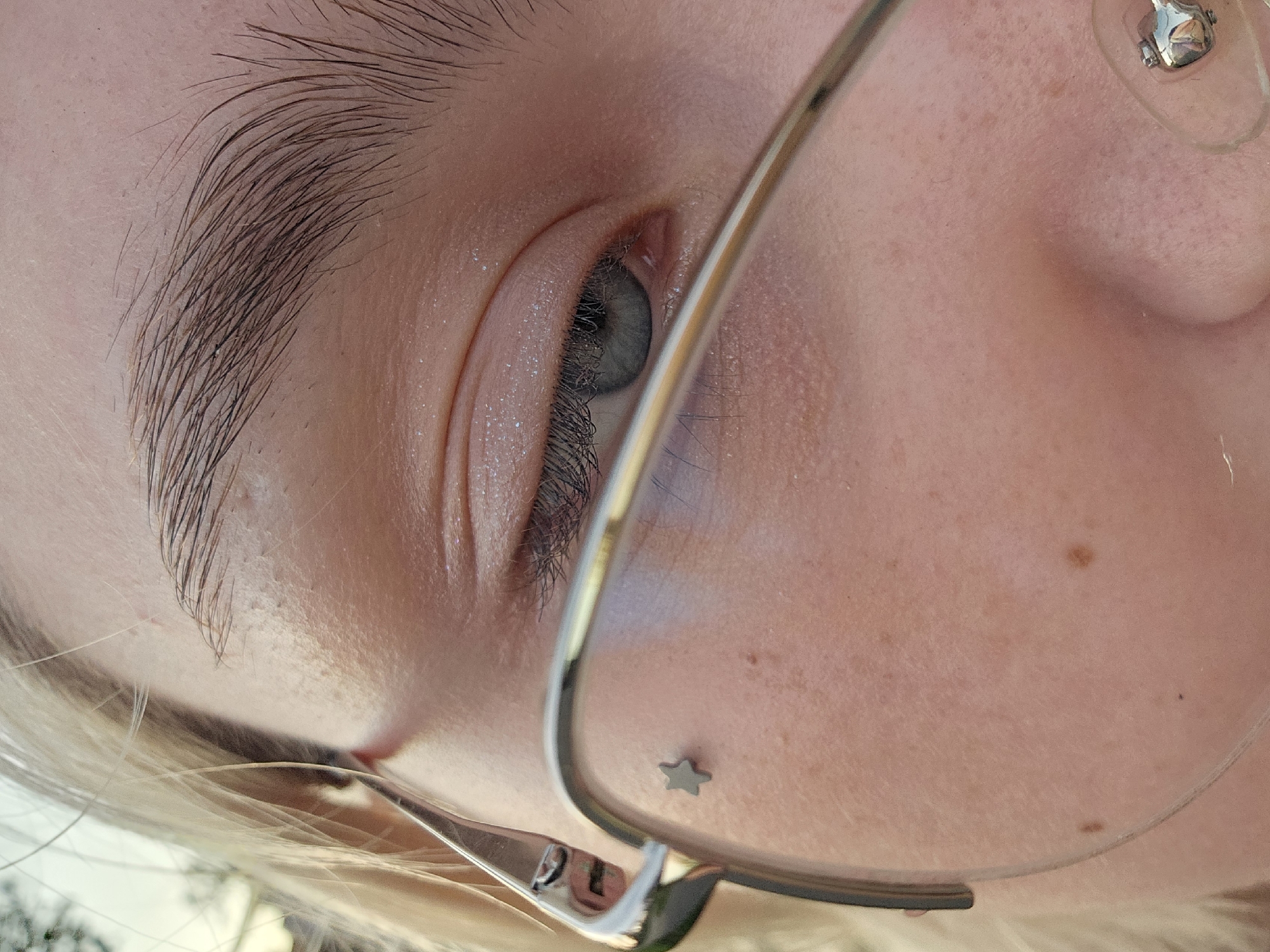 The image is a close-up, eye-level, outdoor shot of a young person's face, with a focus on their left eye and surrounding features. The person has fair skin, light blonde hair, and appears to be wearing glasses with thin, metallic frames. A small, metallic star-shaped piercing is visible on their cheekbone, just below the glasses. The person's eye is light blue or gray and partially visible through the glasses. There is a subtle shimmer of glitter or highlighter on their eyelid. The background is softly blurred, suggesting an outdoor setting with natural light, possibly during the day. The overall scene is intimate and personal, highlighting the details of the individual's face. The image does not contain any information that would indicate a location in Nizhny Novgorod, Russia.