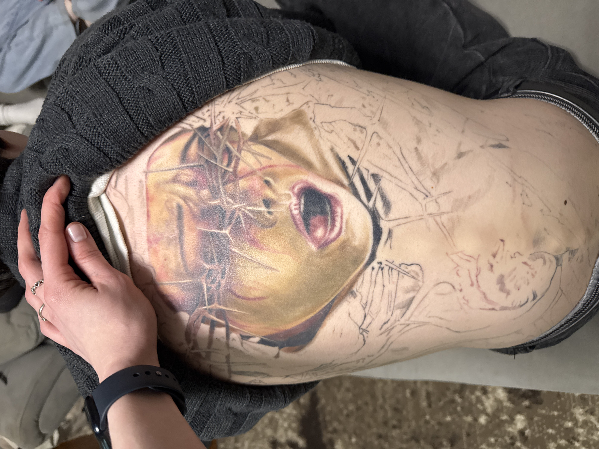 A person with fair skin lies on a light-colored surface, possibly a bed or examination table, and displays a large tattoo in progress on their torso. The tattoo features a dramatic image of a screaming face surrounded by what appears to be a crown of thorns or abstract barbed wire. The face is rendered in detailed color, while the surrounding elements are primarily outlined in black ink, suggesting the tattoo is unfinished. The person wears a dark gray, knitted garment and possibly dark denim jeans. A hand with a black watch band is placed on the torso near the tattoo. The location is the city of Perm, Russia.