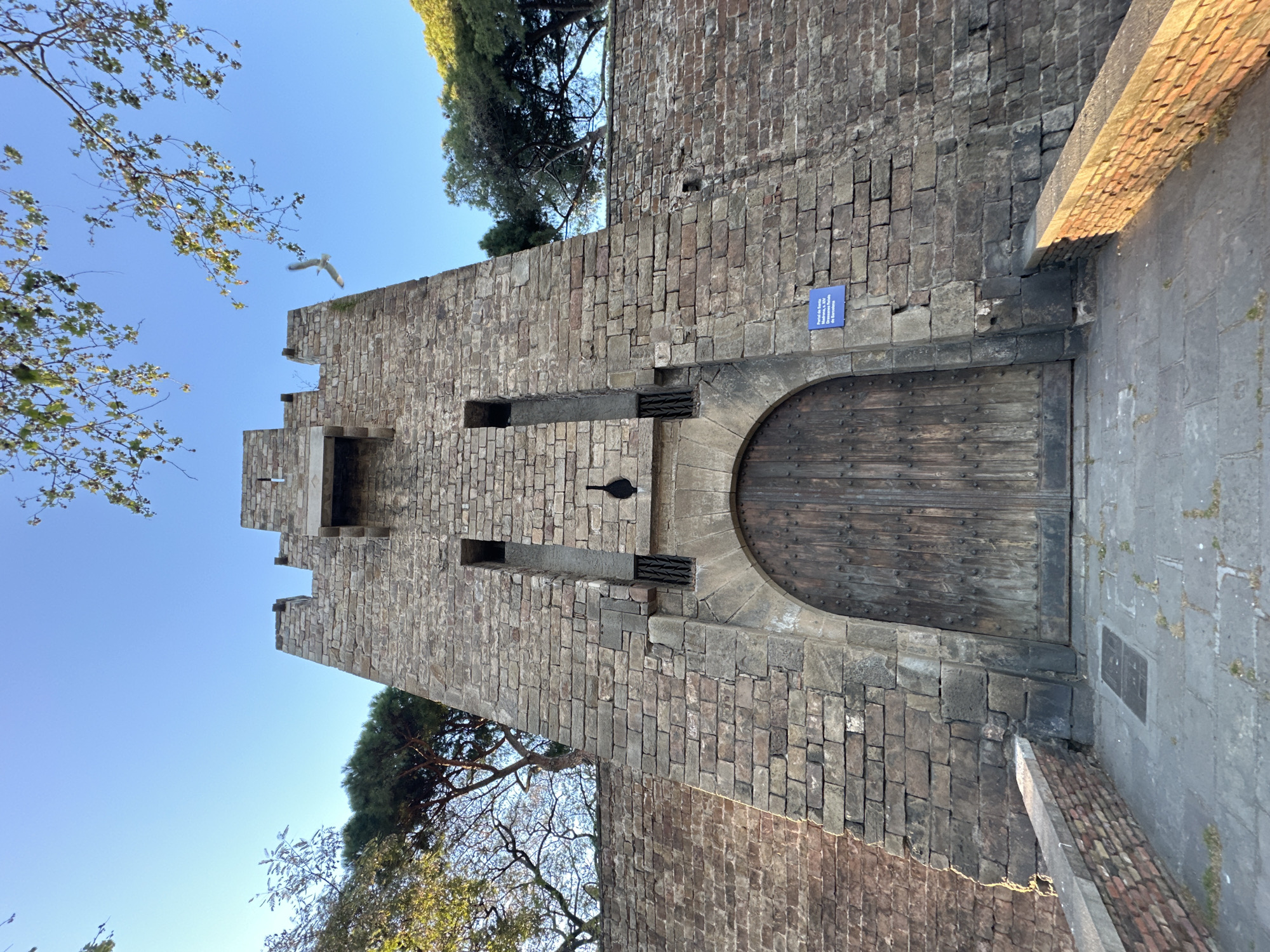 The image shows a stone tower with a wooden door, part of Montjuïc Castle in Barcelona, Spain. The tower is made of light brown stone bricks, with battlements visible at the top. There is a small opening in the tower's wall, with an iron grating below it. A wooden door, arched at the top, is set into the base of the tower. A blue informational sign is affixed to the wall next to the door. Trees and a blue sky are visible behind and around the tower. A bird flies in the sky above the tower.