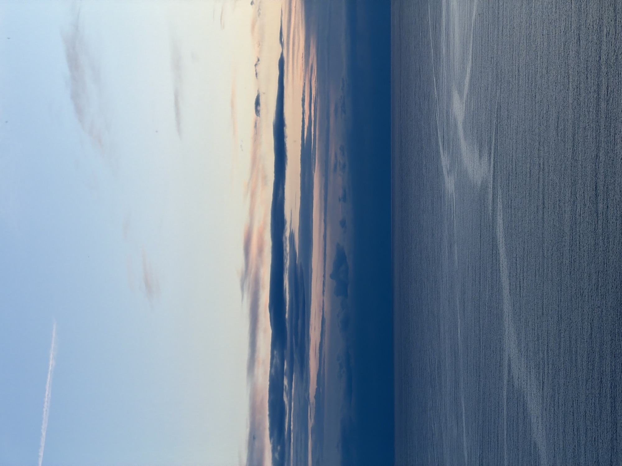 This photograph depicts a view of the Adriatic Sea, likely near Budva, Montenegro, at what appears to be either sunrise or sunset. The sky transitions from a light blue to a darker hue, with bands of clouds tinged with pink and peach colors. A contrail stretches across the lighter part of the sky. The sea, which occupies the right side of the image, is a dark blue with subtle patterns of waves or reflections.