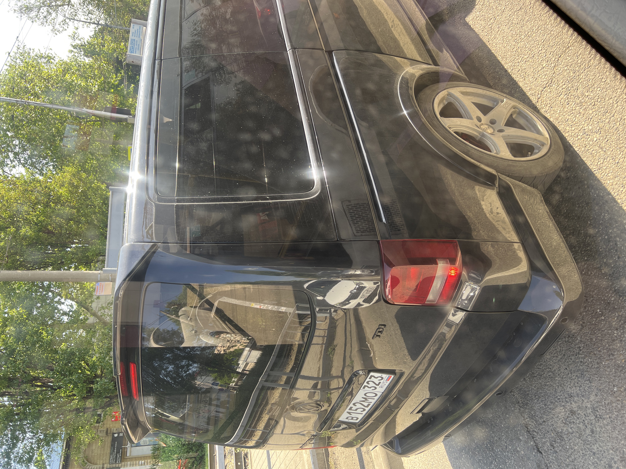 A dark-colored van, likely a Volkswagen Transporter or similar model, is parked on a street. The rear of the vehicle is prominently displayed, showing a "TDI" badge and a Russian license plate reading "B152MO323 RUS." The vehicle appears to be in Krasnodar, Russia, as evidenced by the "RUS" code on the license plate. The rear of the van reflects street elements, including buildings and potentially other vehicles. The van's wheel is visible. Trees are visible in the background. The day appears bright and sunny.